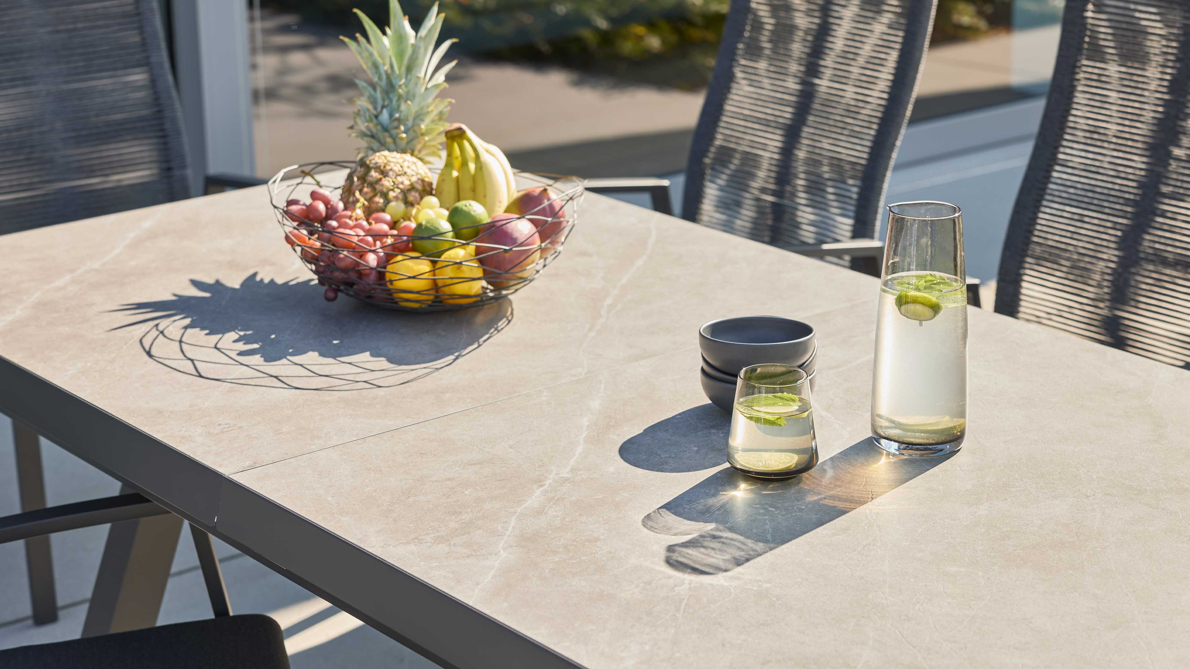 Rechteckiger Tisch mit hellgrauer Steinoberfläche und Metallrahmen, darauf ein Drahtkorb mit Obst, Karaffe und Glas mit Limette.