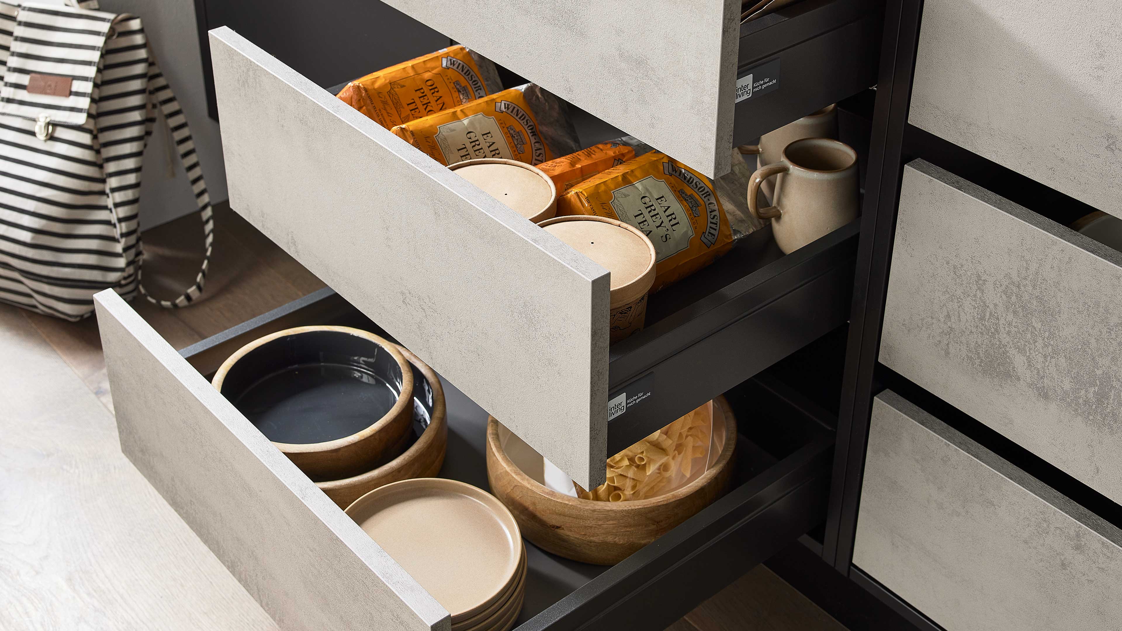 Geöffnete hellgraue Küchenschubladen: oben Teepackungen, Dosen und Tassen; unten Teller, Holzschalen und Pasta, links Rucksack.