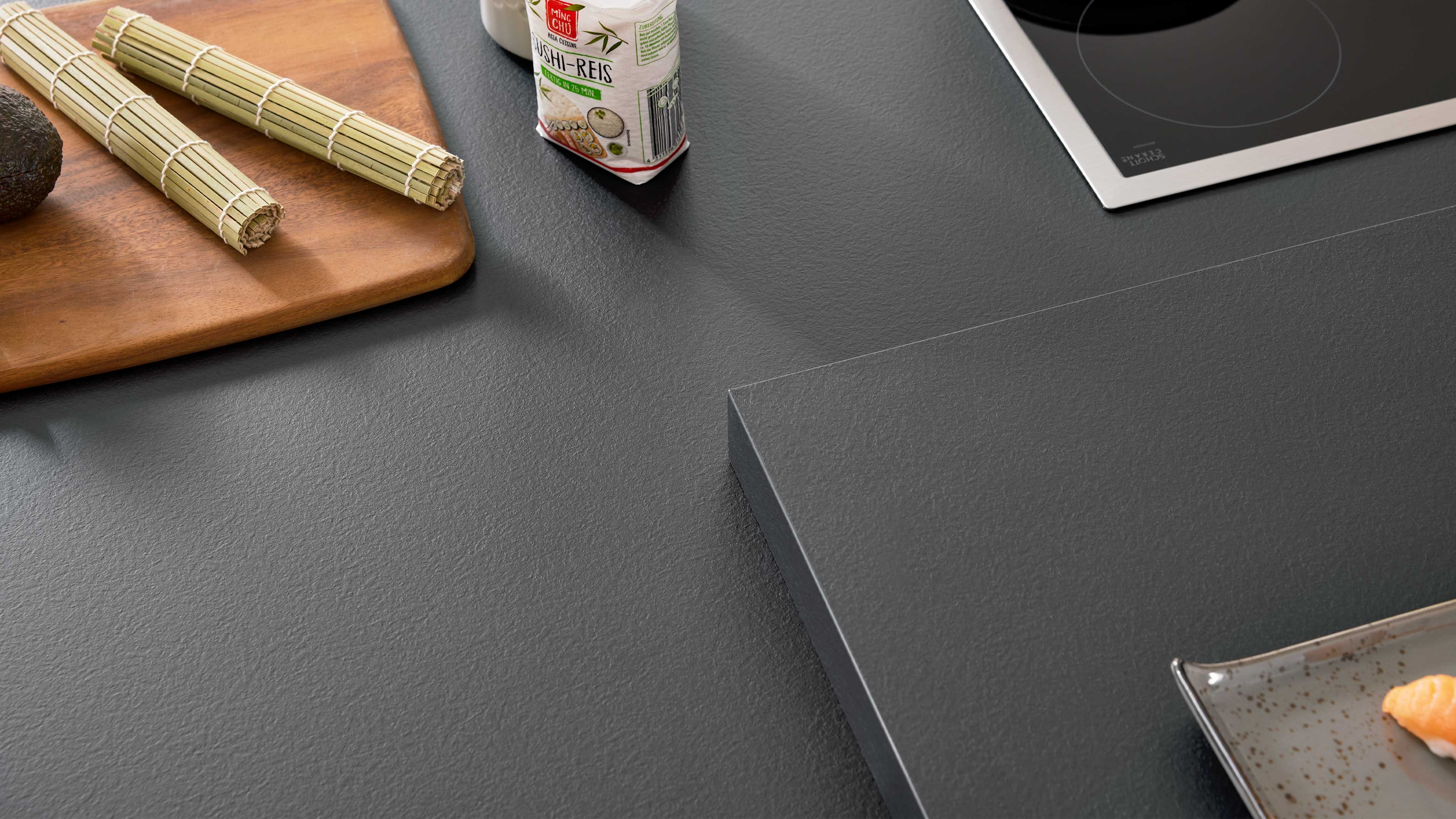 Dunkle Küchenarbeitsplatte mit Kochfeld oben rechts, Holzbrett mit Bambusmatten und Avocado links, Reispackung und Teller mit Lachs unten.