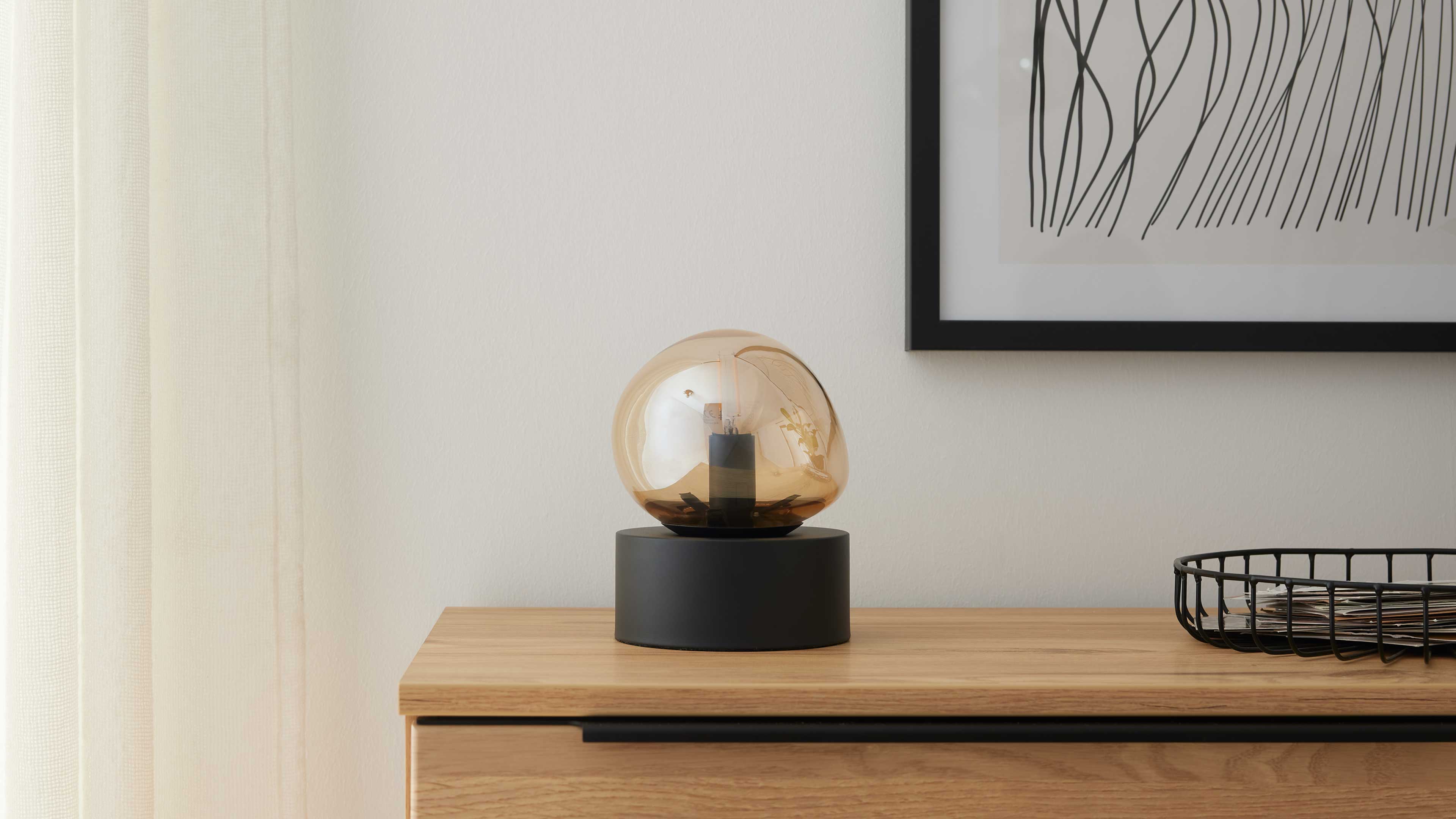 Auf einer Holzkommode steht eine Tischlampe mit bernsteinfarbener Glaskugel auf schwarzem Zylinderfuß, rechts ein Drahtkorb.