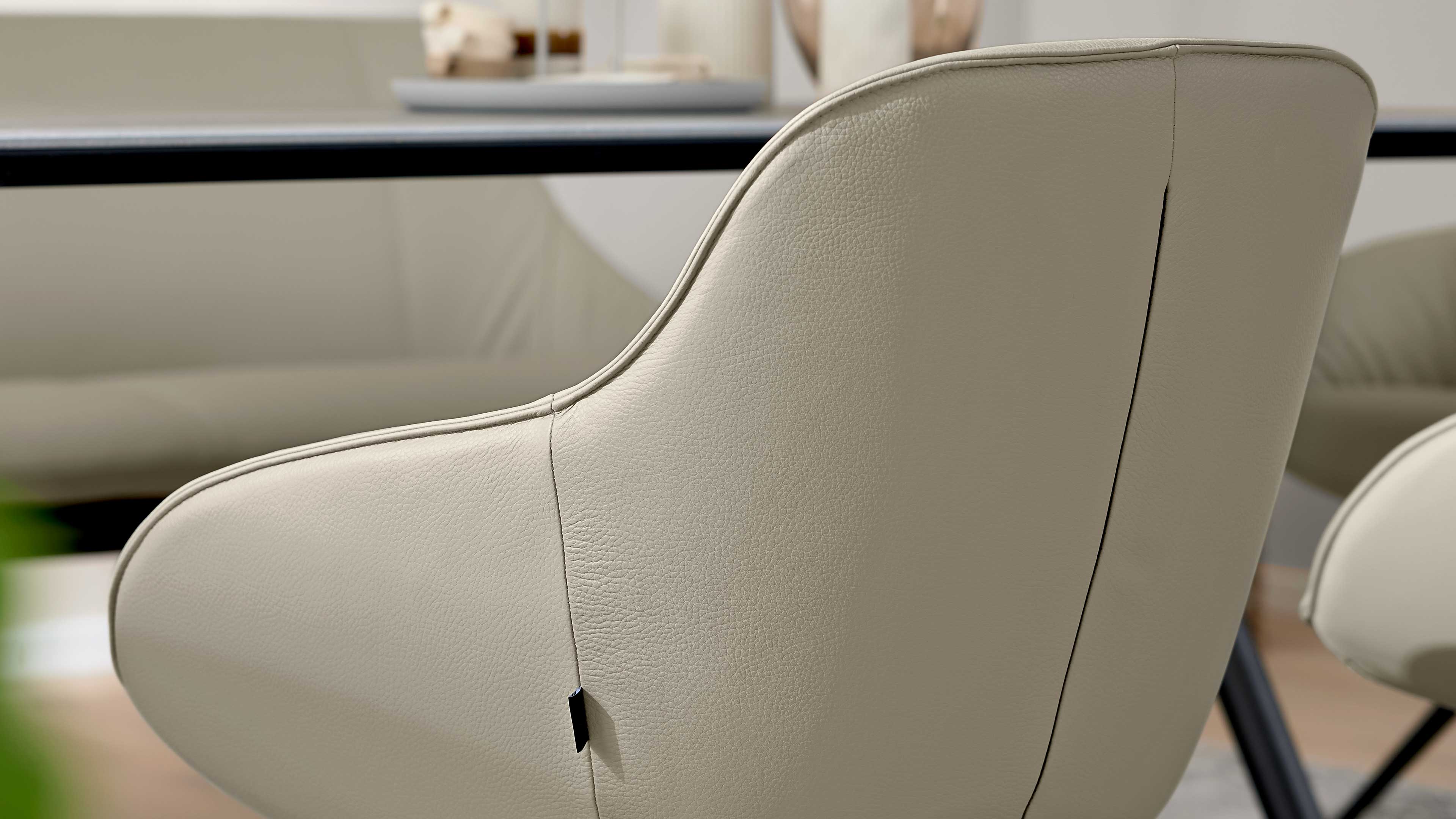 Beige Kunstlederstuhl mit geschwungener Rückenlehne, sichtbaren Nähten und kleinem schwarzen Stofflabel, vor Tischkante.