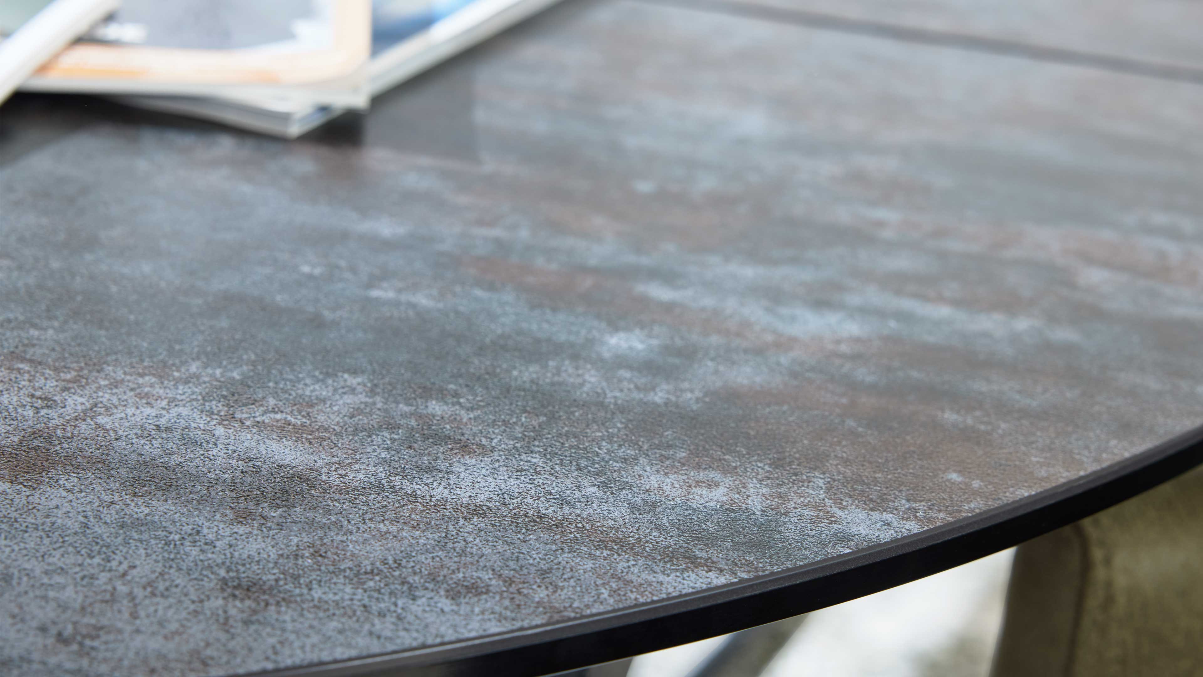 Runder Tisch mit dunkelgrauer, leicht fleckiger Oberfläche, sichtbarer schwarzer Kante und oben links unscharfem Zeitschriftenstapel.