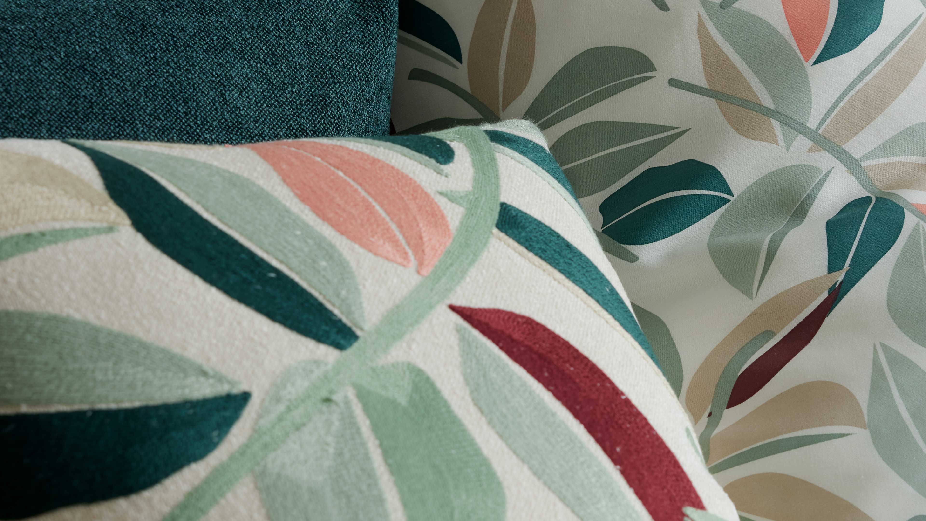 Kissen mit Blattmuster in Grün, Türkis, Koralle und Dunkelrot vor grünem Polster und heller Tapete mit Blättern.
