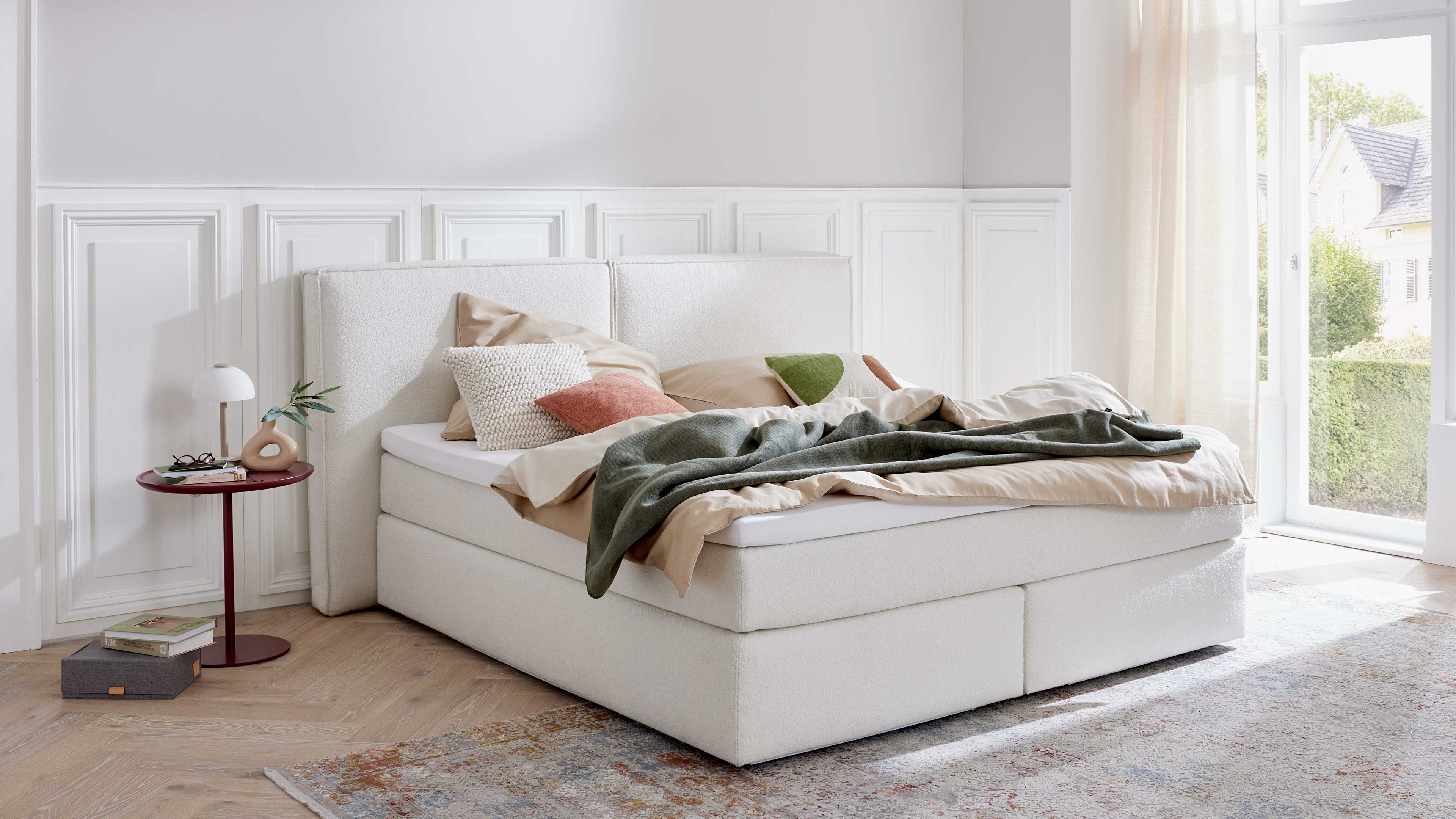 Weißes Polsterbett mit hohem Kopfteil, beiger Bettwäsche, mehreren Kissen und grünem Überwurf; daneben runder roter Nachttisch mit Lampe.