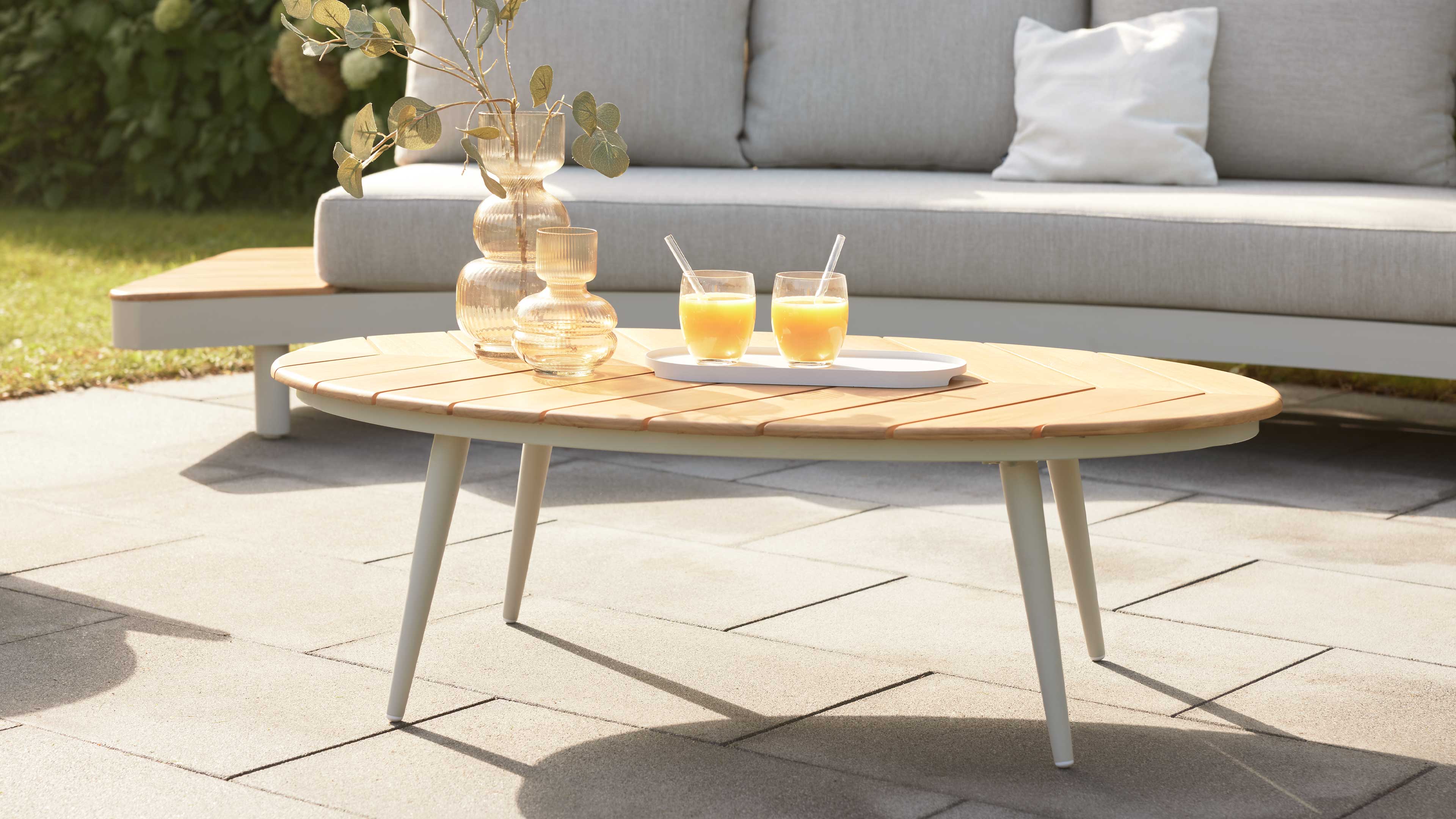 Ovaler niedriger Couchtisch mit hellholzplatte und weißen Metallbeinen, darauf zwei Gläser Orangensaft, eine Schale und zwei Glasvasen.