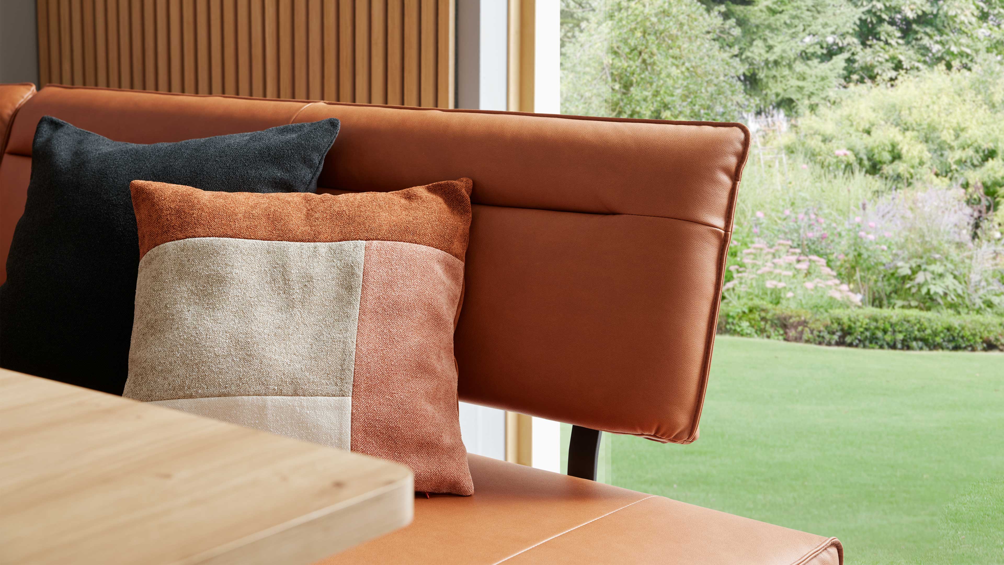 Braune Lederbank mit vorderem beige-rostfarbenem Patchworkkissen und dunklem Kissen dahinter, links Tischkante, rechts Garten.