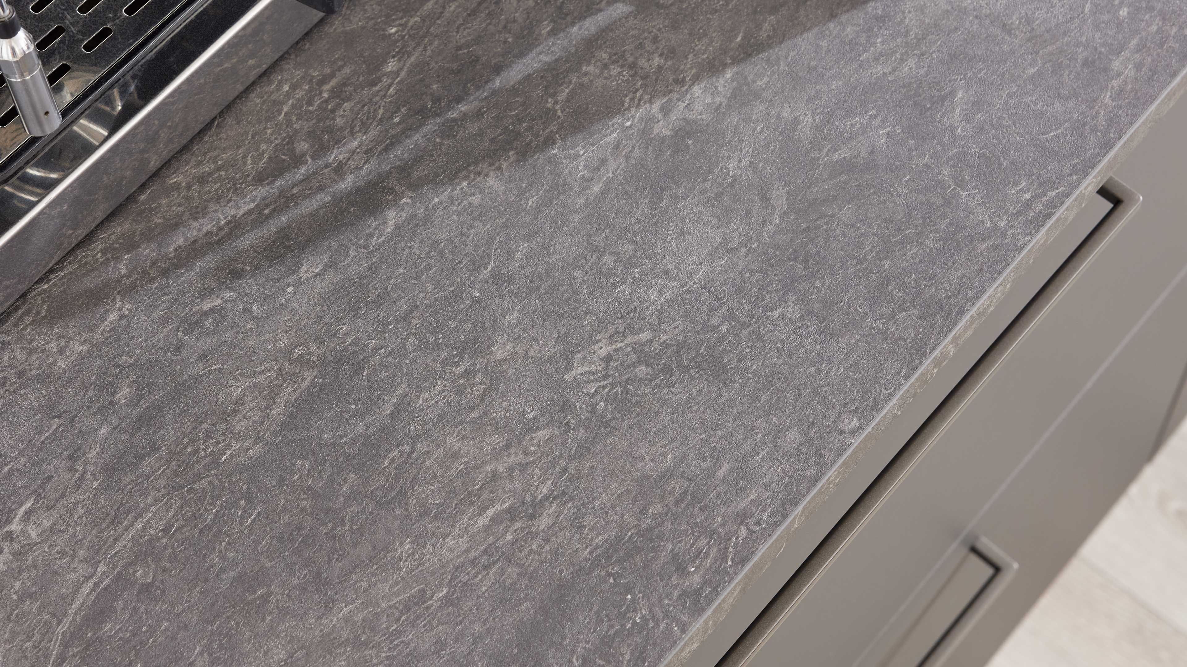 Graue strukturierte Arbeitsplatte in Steinoptik, links ein Teil einer Edelstahl-Abtropfschale, rechts graue Schubladenfront mit Griffmulde.