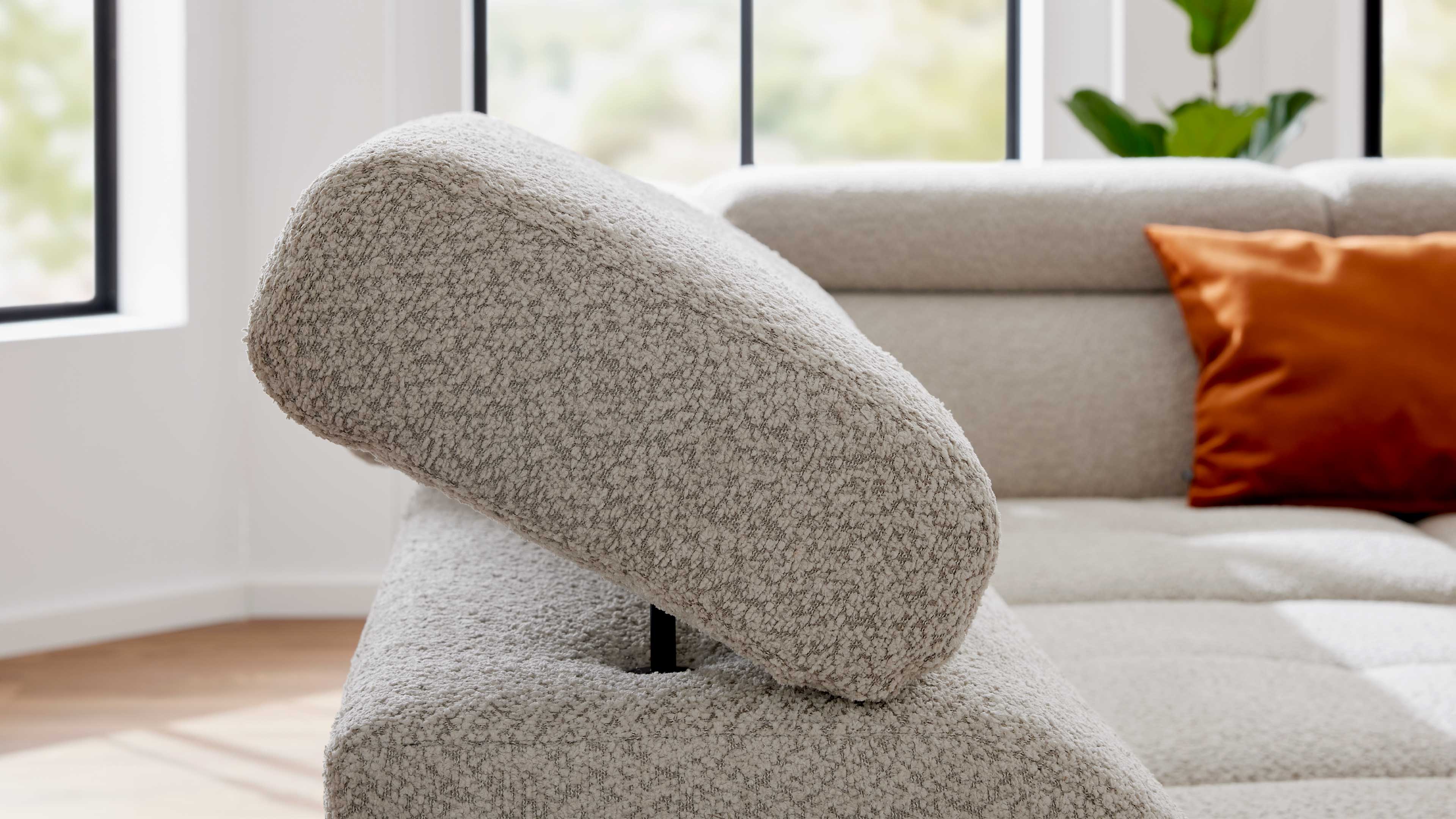 Nahaufnahme einer hellgrauen, boucleartigen Kopfstütze eines Sofas; im Hintergrund ein orangefarbenes Zierkissen und Fenster.