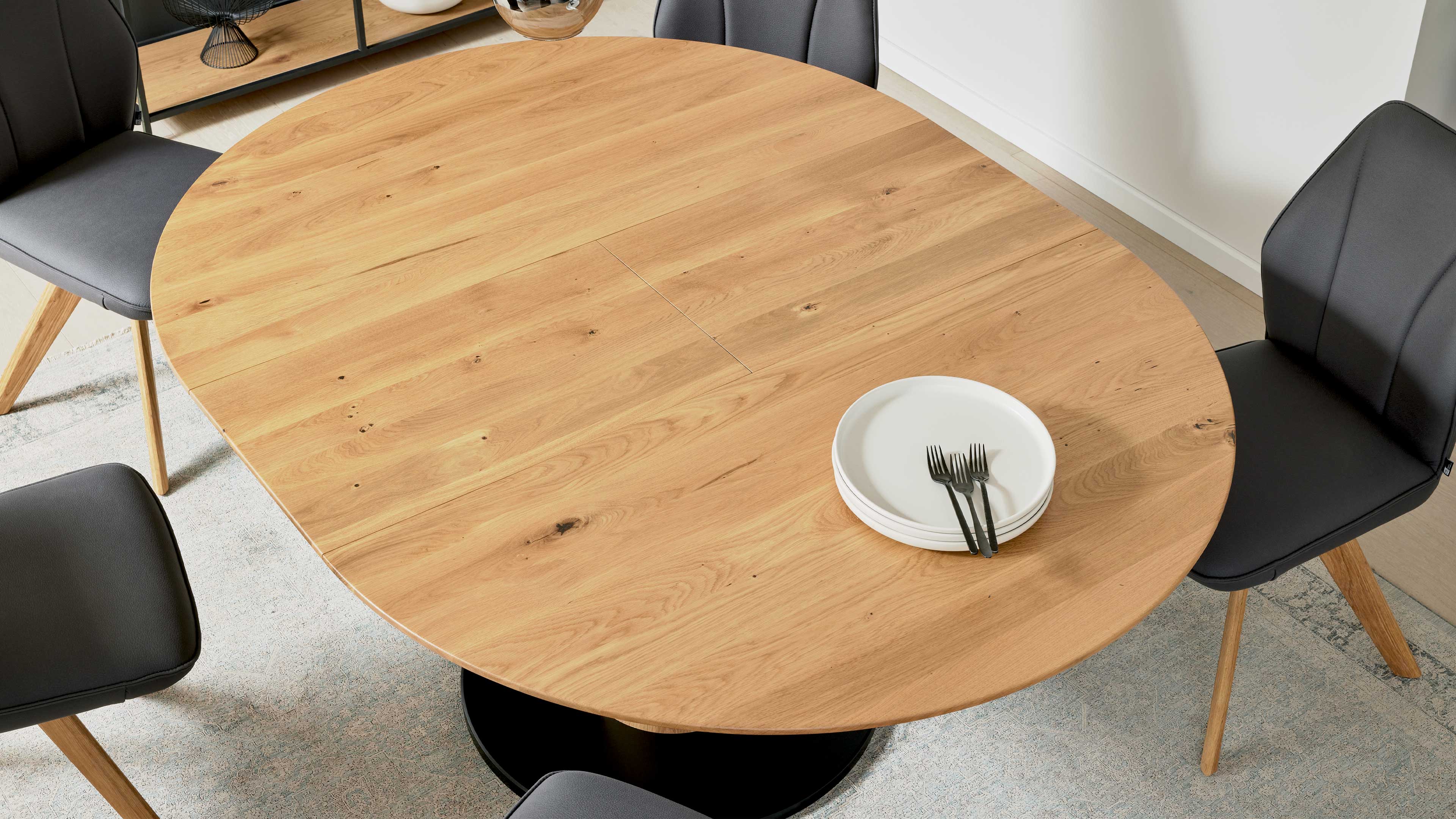Runder hellbrauner Holz-Esstisch mit mittiger Fuge, rechts gestapelte weiße Teller mit vier Gabeln und dunkle Polsterstühle.