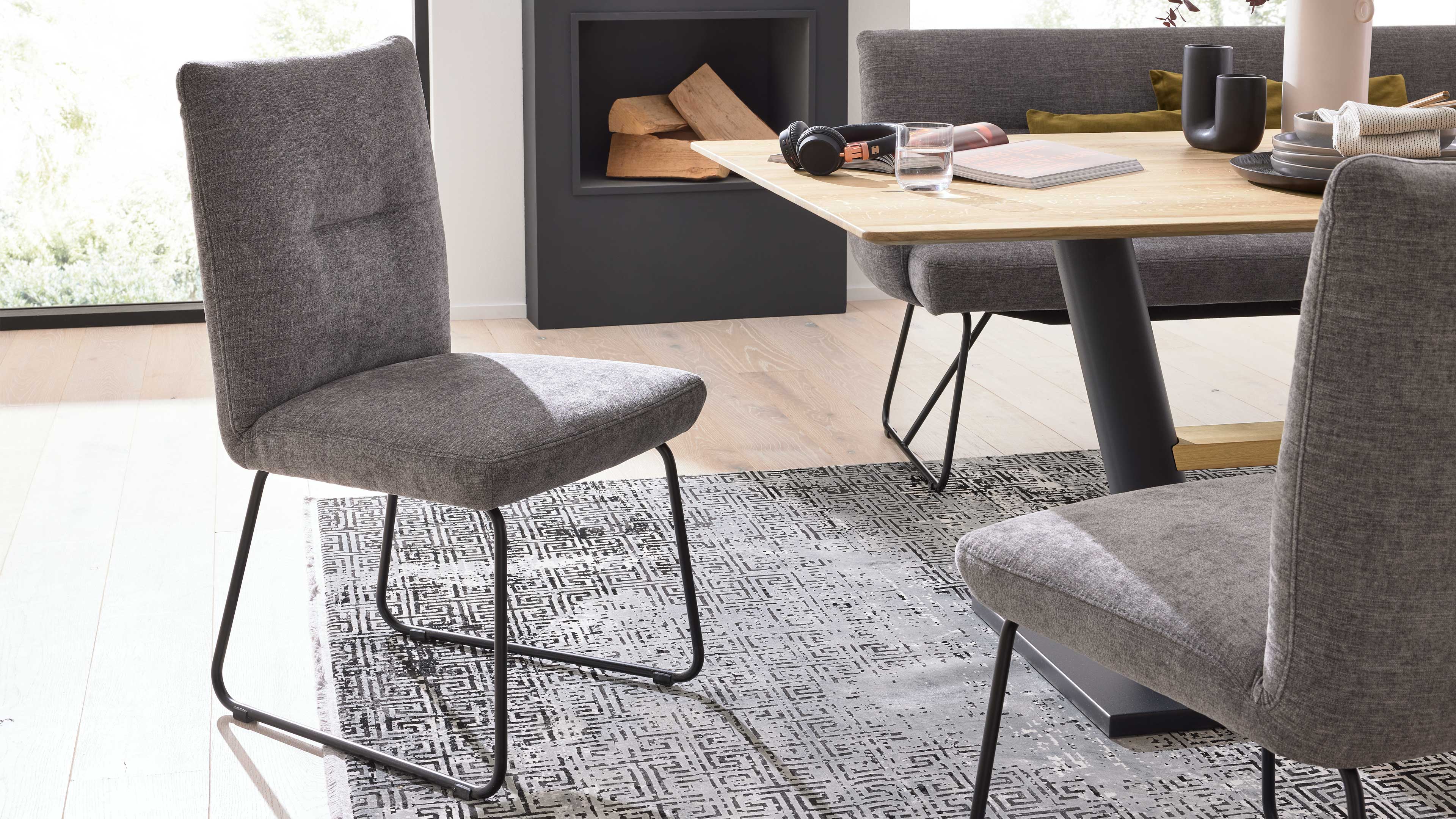 Grau gepolsterter Stuhl mit schwarzer Metallkufe auf gemustertem Teppich neben Holztisch und Bank mit Kissen, Kamin mit Feuerholz.