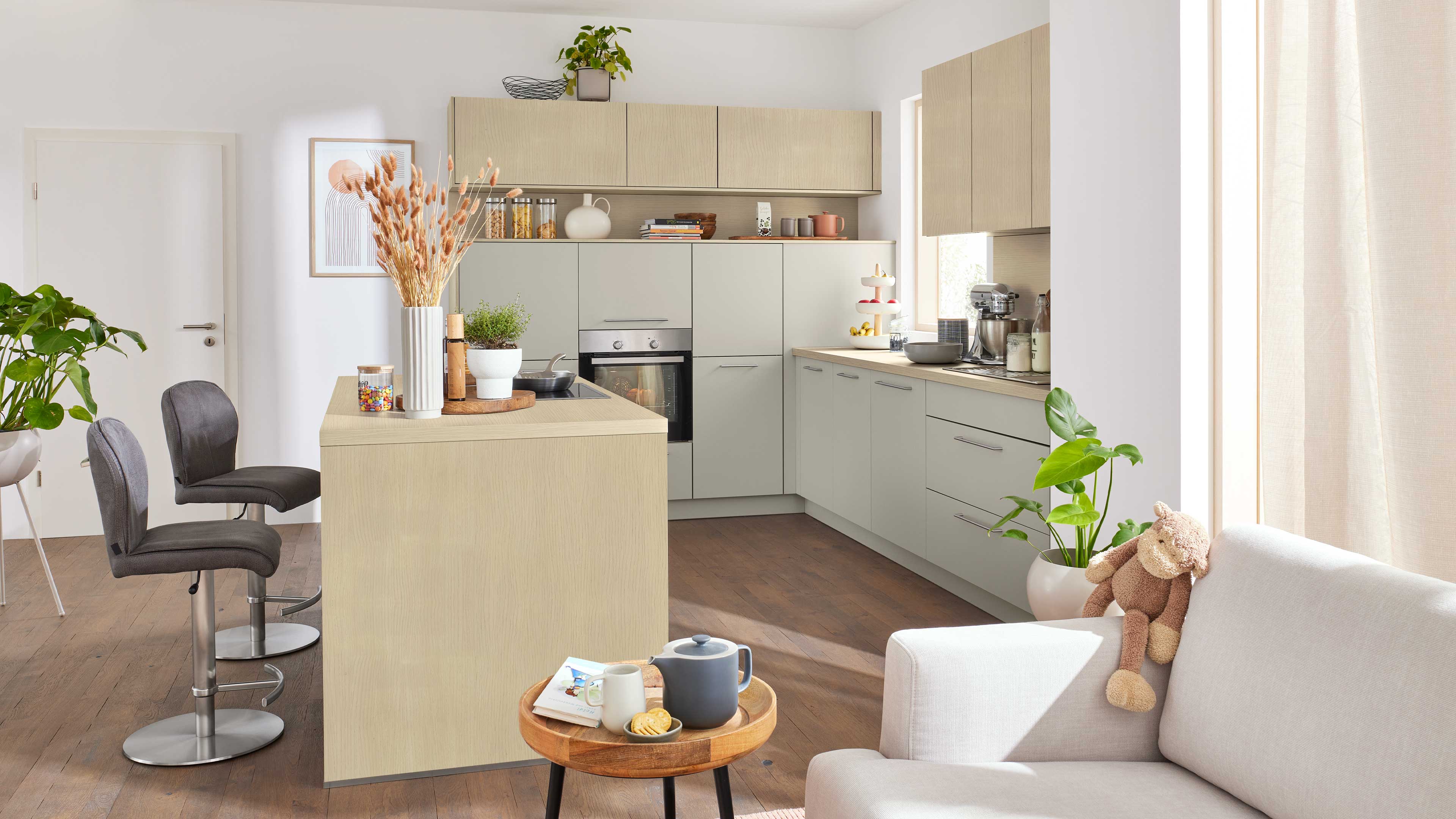 Helle Küchenzeile mit Holz- und grauen Fronten, Insel mit Kochfeld und Backofen, zwei graue Barhocker, Pflanzen und Sofa mit Teddybär.