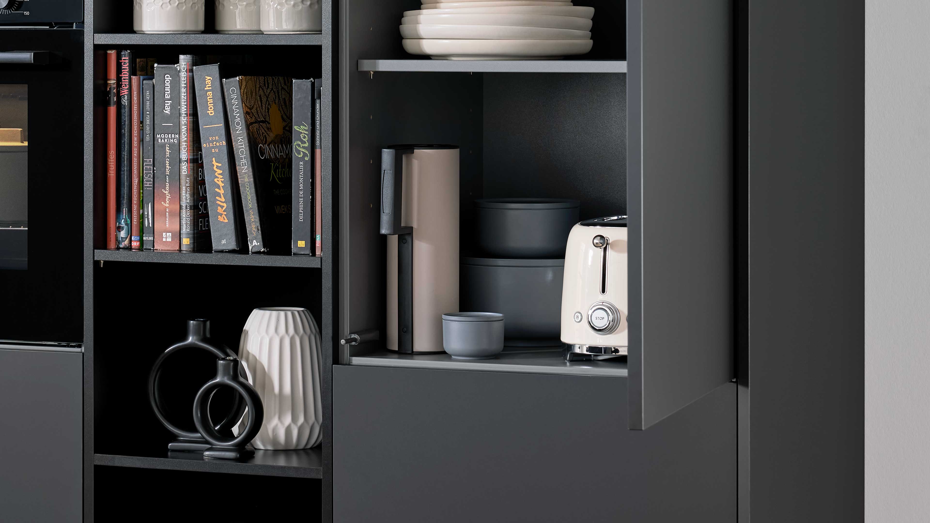 Offenes Fach eines dunkelgrauen Küchenschranks mit beigem Toaster rechts, heller Kanne, grauen Schalen und Tellern, links Bücher und Vasen.