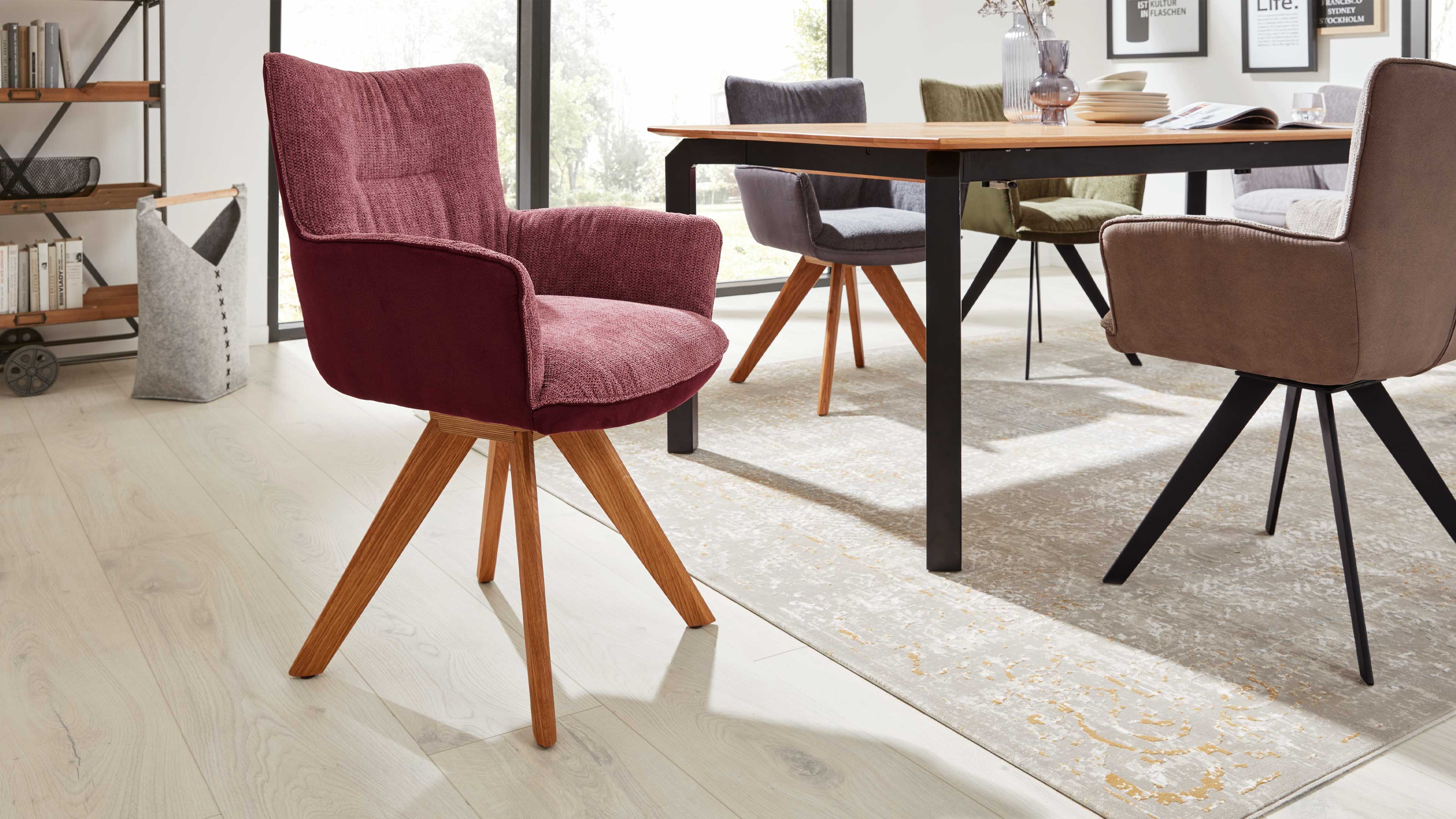 Burgunderfarbener, gepolsterter Stuhl mit vier nach außen geneigten Holzbeinen im Vordergrund, daneben Esstisch und weitere Stühle.