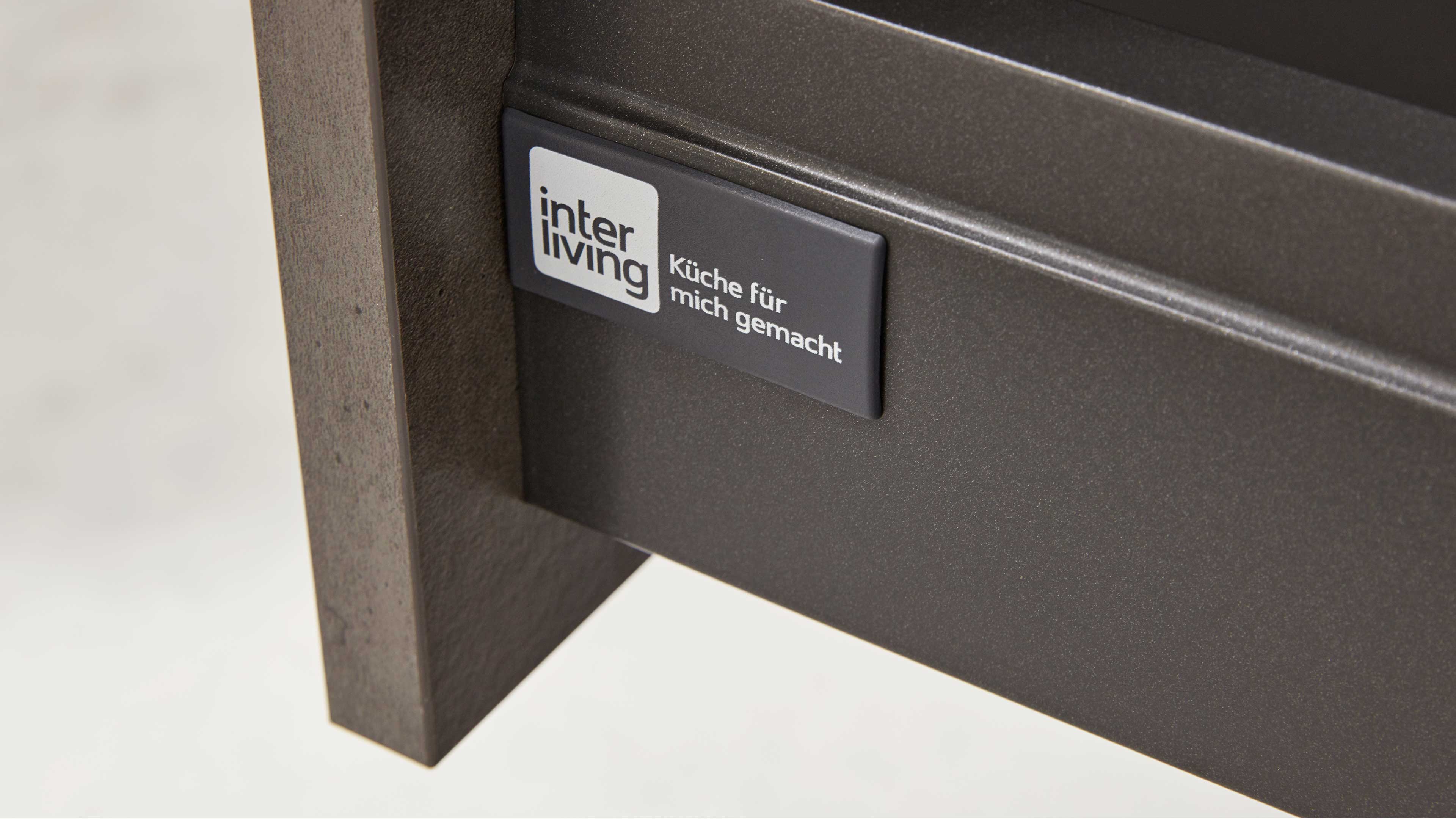 Nahaufnahme einer dunkelgrauen Schubladenfront mit rechteckigem Aufkleber 'inter living – Küche für mich gemacht' und sichtbarer Holzstütze.