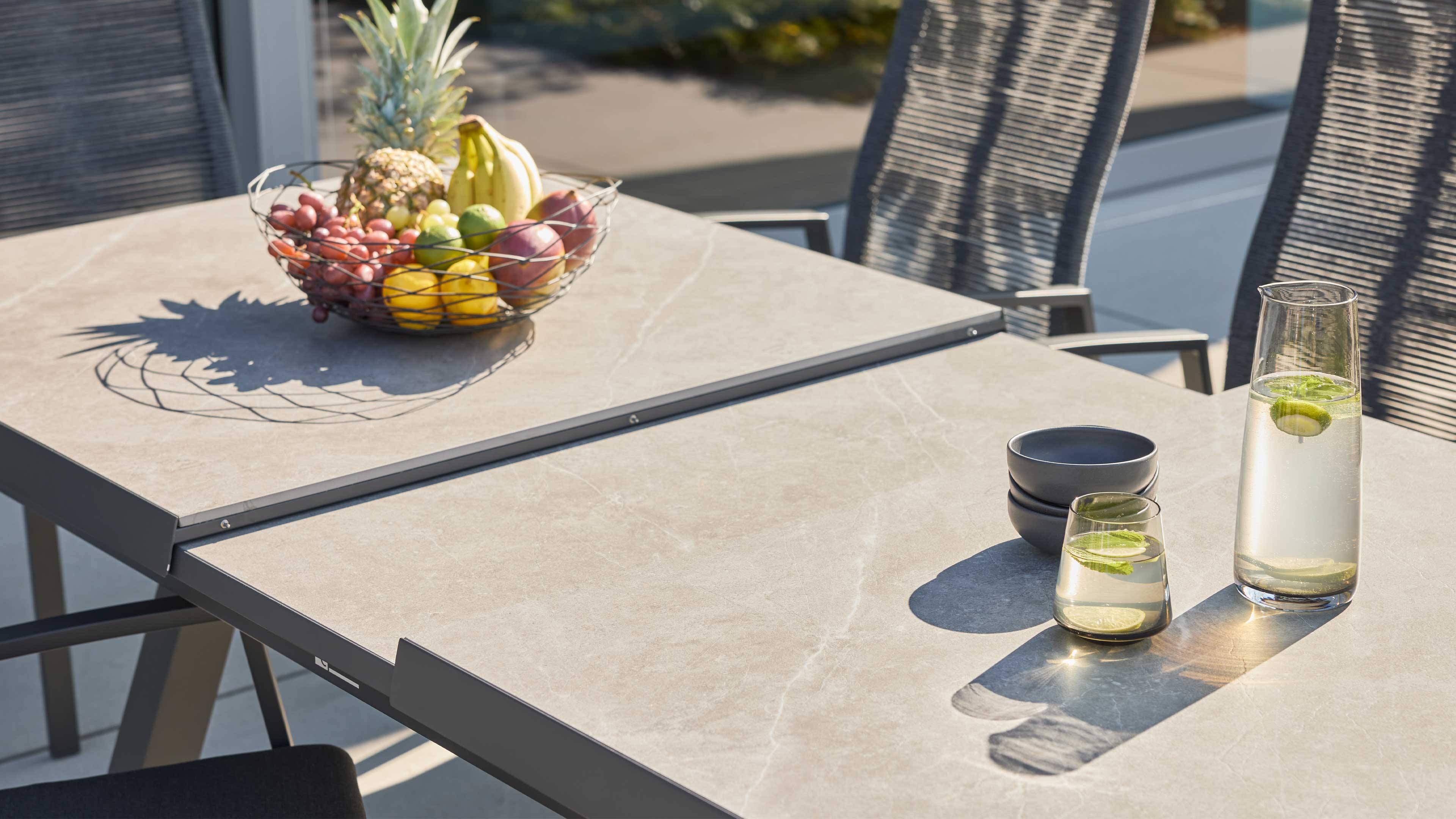 Großer rechteckiger Gartentisch mit heller Stein-Optik, Drahtkorb mit Früchten links, rechts Karaffe und Glas mit Limettenscheiben.