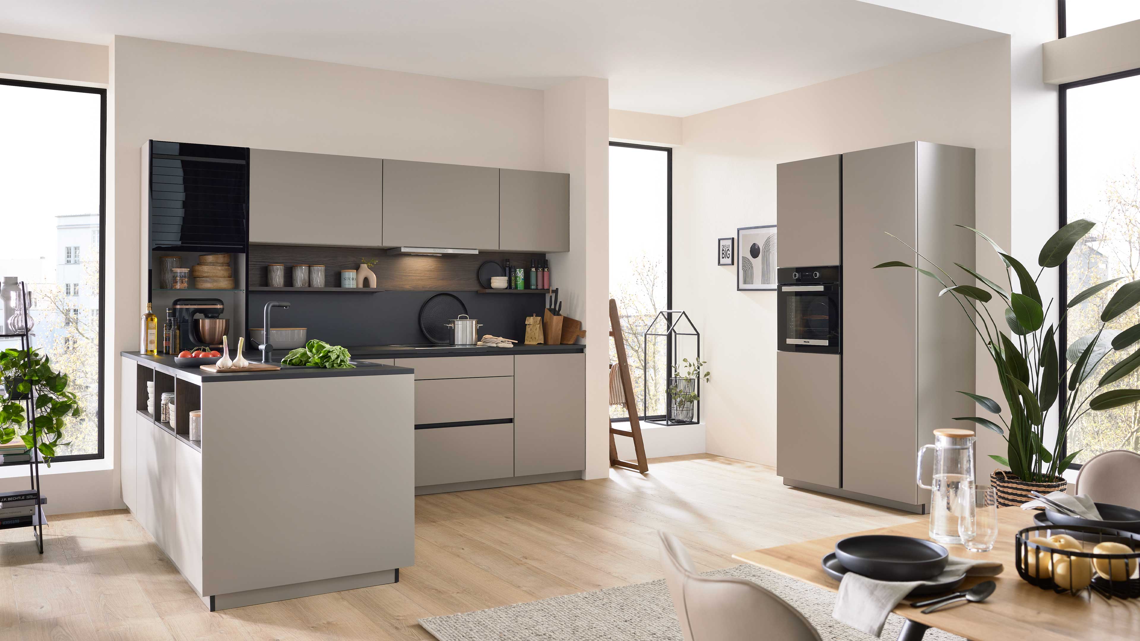 Moderne Küche mit hellgrauer Küchenzeile und Kochinsel, schwarzer Arbeitsplatte, Hochschrank mit Backofen und Pflanzen.