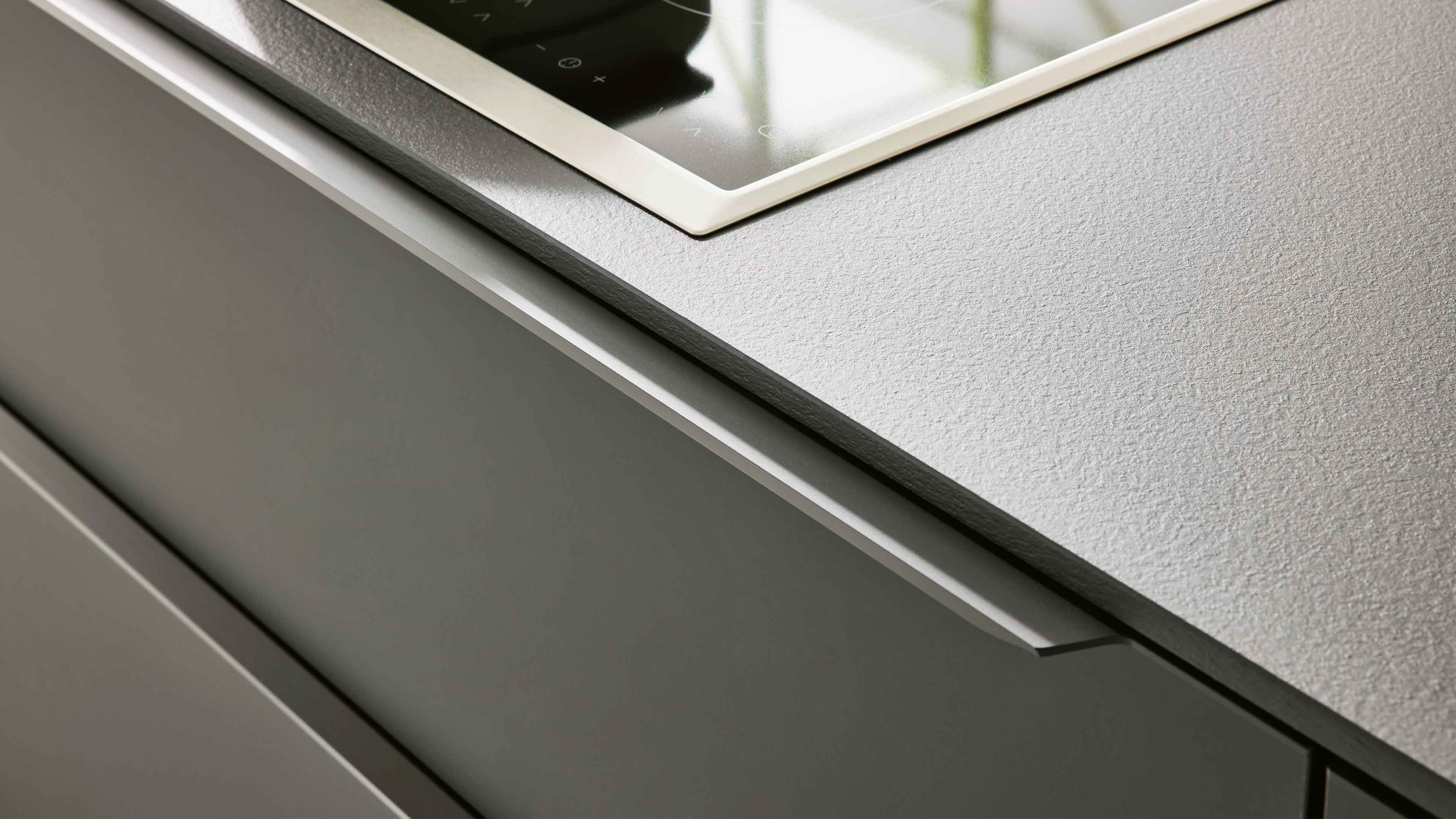Naheinstellung einer hellgrauen, strukturierten Arbeitsplatte mit eingelassenem schwarzen Kochfeld und dunkelgrauer Schublade mit Griffleiste.