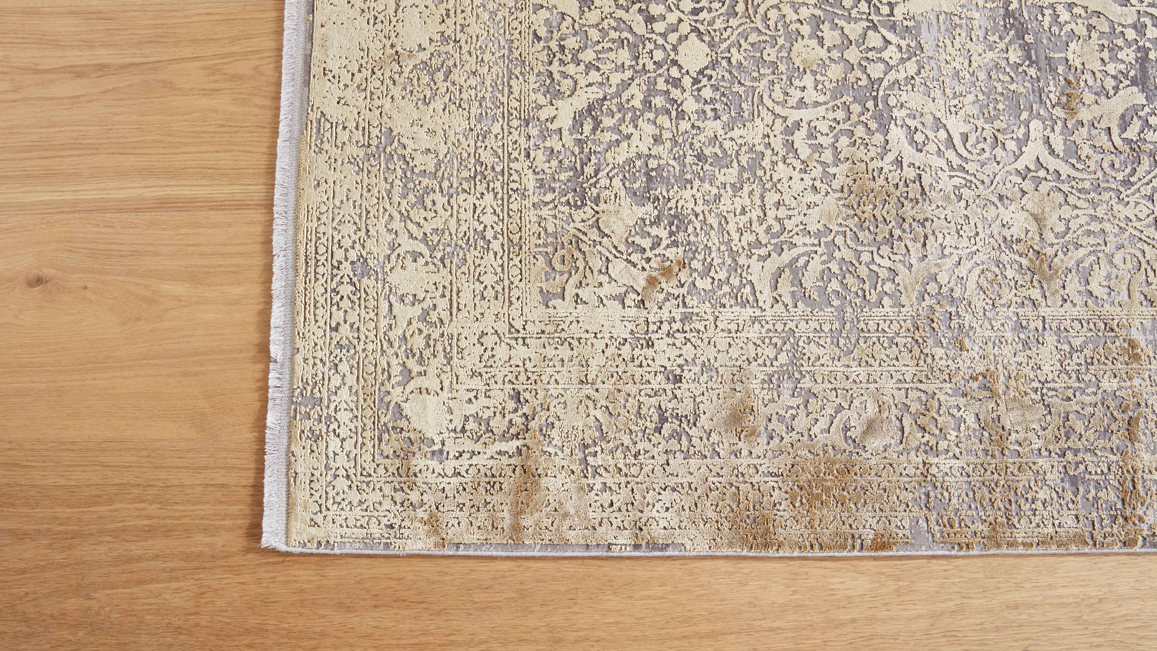 Teppich mit hellbeigem, floralen und geometrischen Ornamenten, linke Fransen sichtbar, liegt auf hellem Holzfußboden.