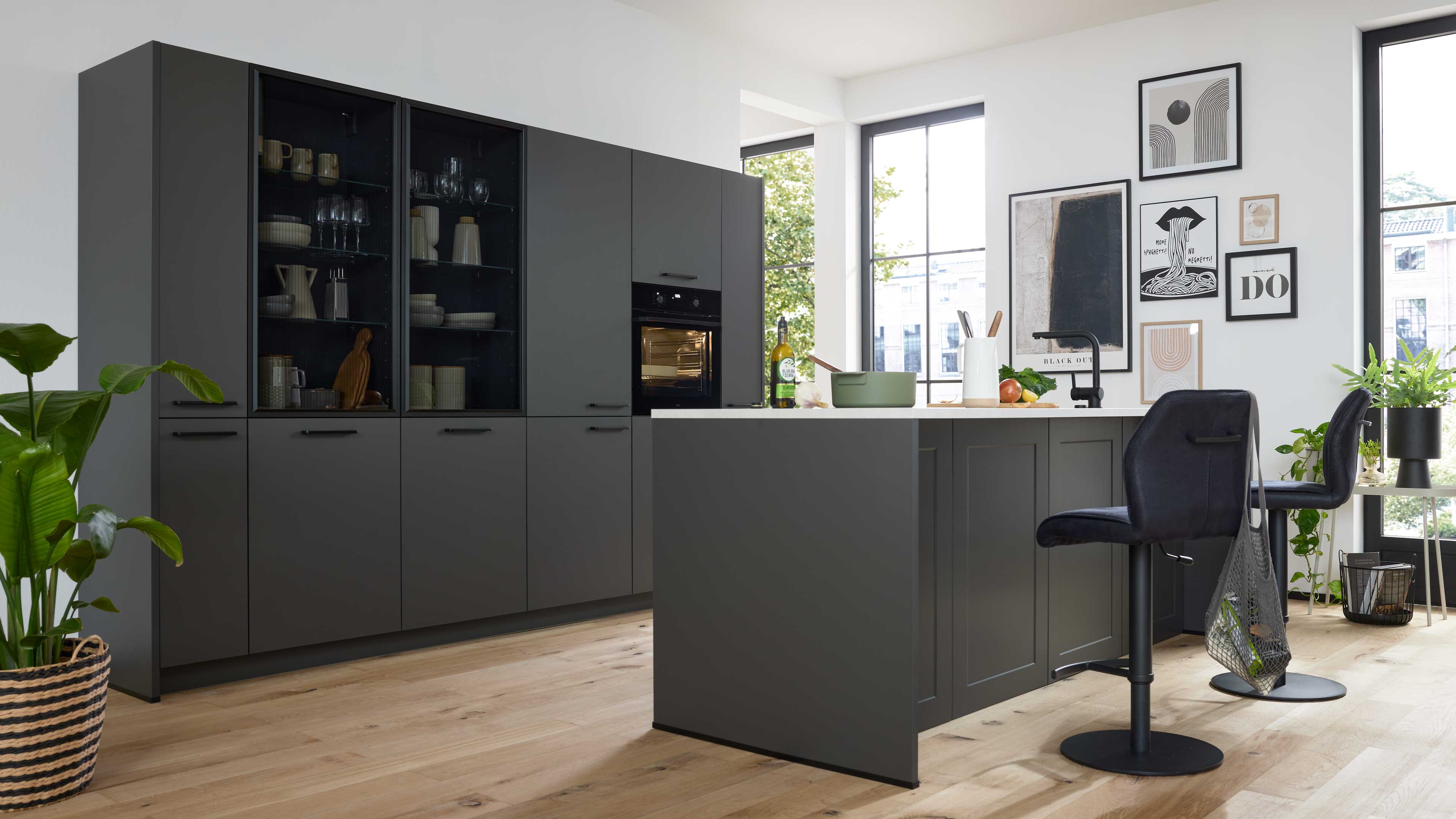 Große dunkelgraue Küchenzeile mit Insel, Glastüren, eingebautem Ofen, Barhockern, Zimmerpflanzen und Bilderwand.