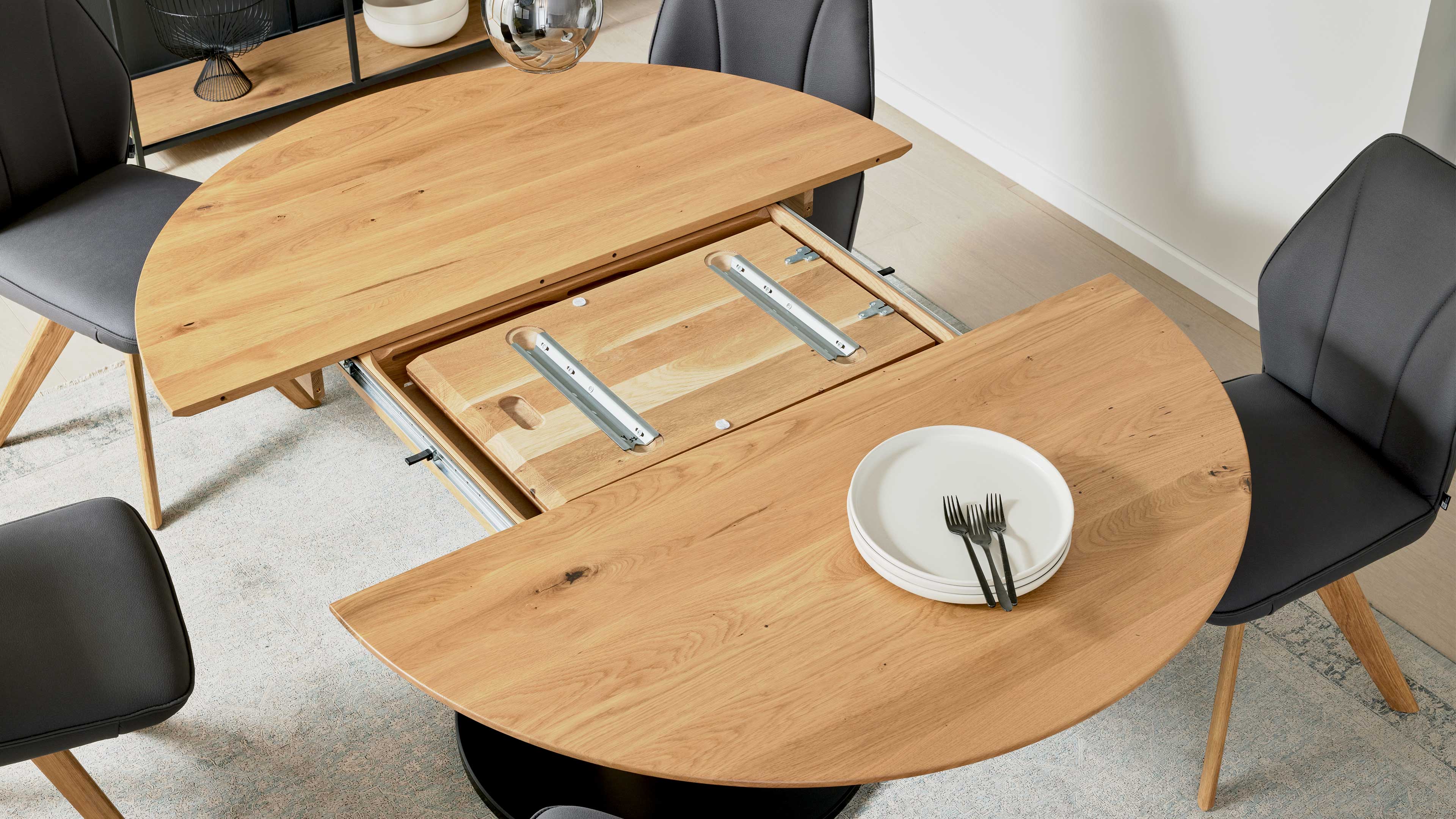 Runder Esstisch mit offener Mittelverlängerung und sichtbaren Metallschienen, dazu drei dunkelgraue Polsterstühle.