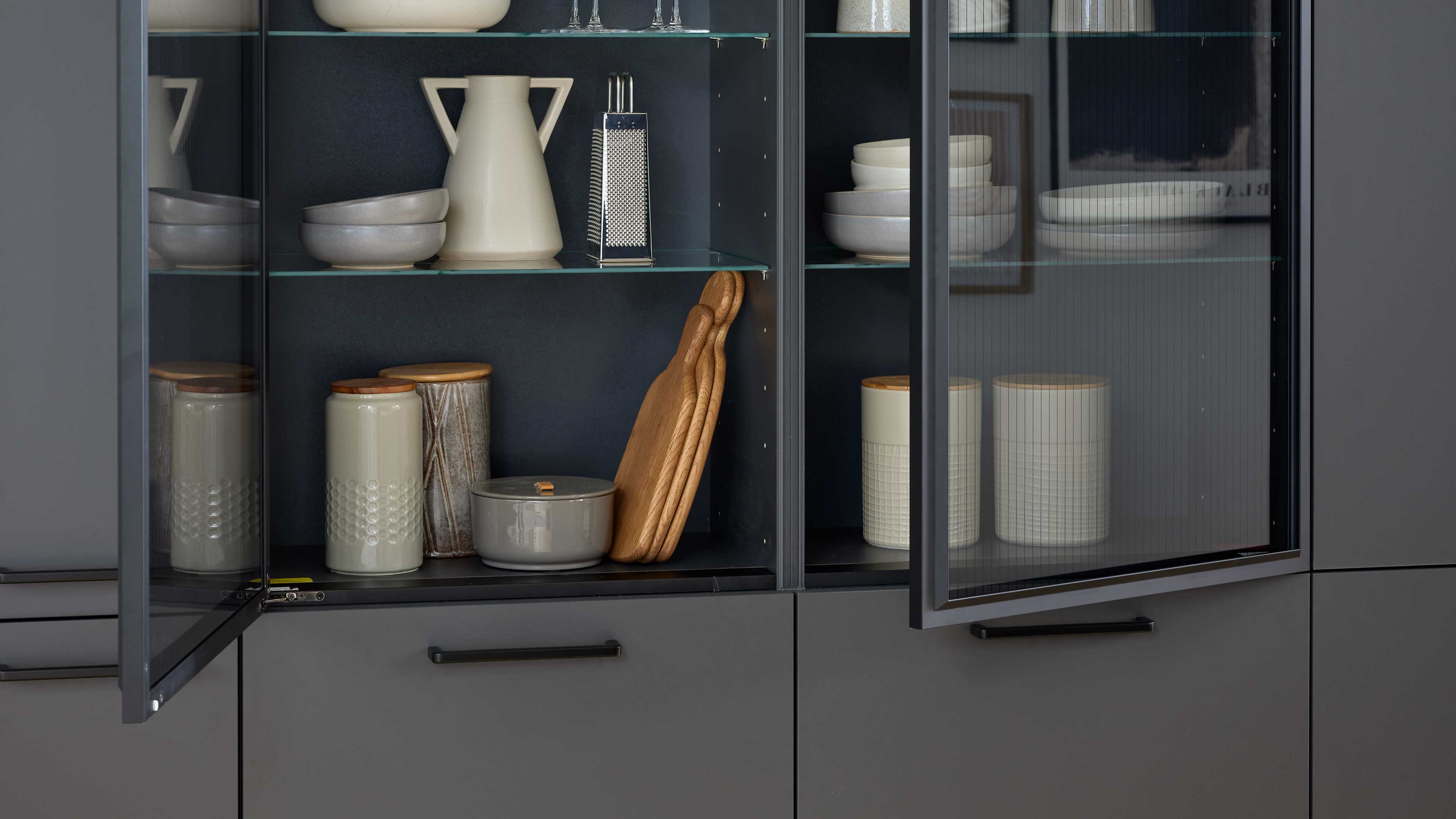Geöffneter grauer Küchenschrank mit Glastüren, innen Keramikdosen, Schalen, ein Topf, Holzschneidebretter und eine Reibe.
