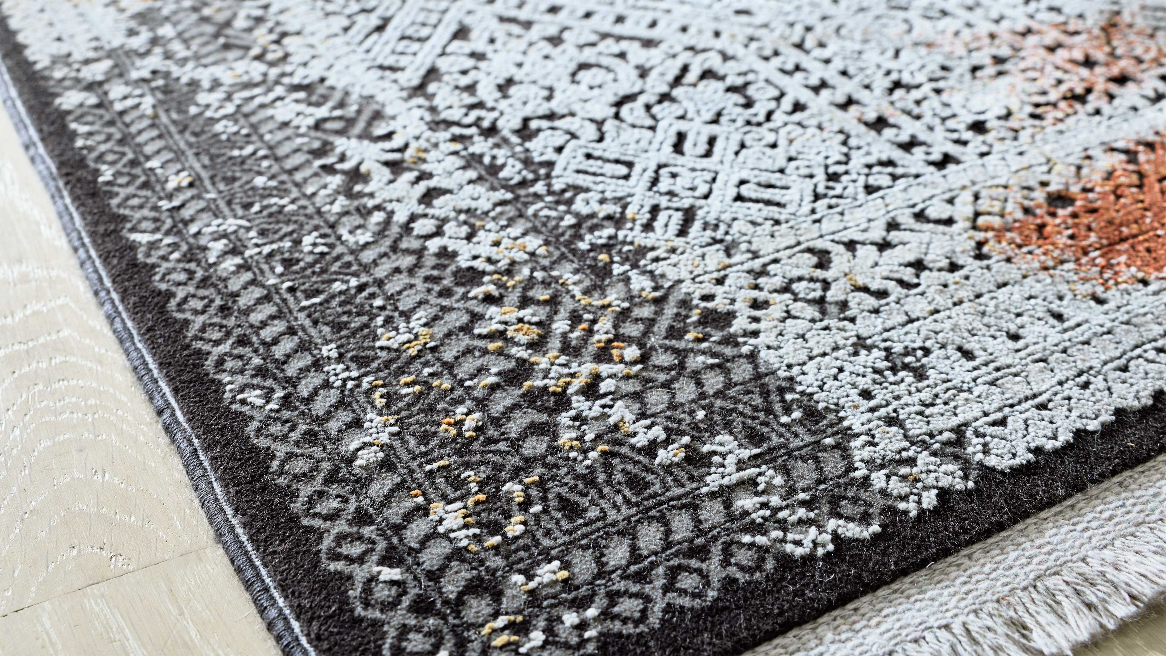 Dunkelgrauer Teppich mit hellgrau-weißer Reliefmusterung und vereinzelten gelben Punkten, Fransen am Rand auf hellem Holzboden.