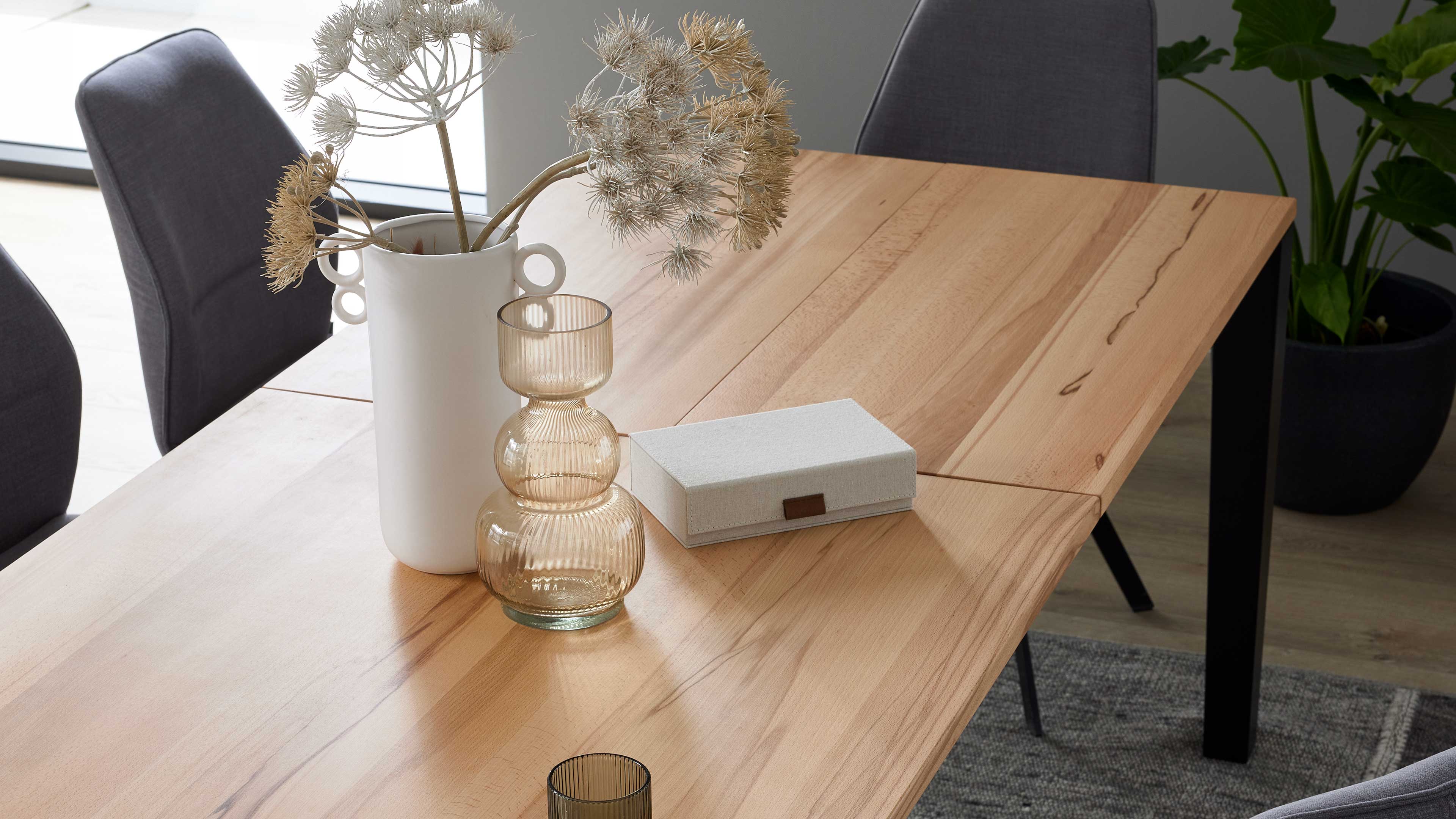 Holztisch mit sichtbarer Maserung, darauf weiße Vase mit getrockneten Dolden, gestufte Glasvase und rechteckige Stoffbox; graue Stühle im Hintergrund.