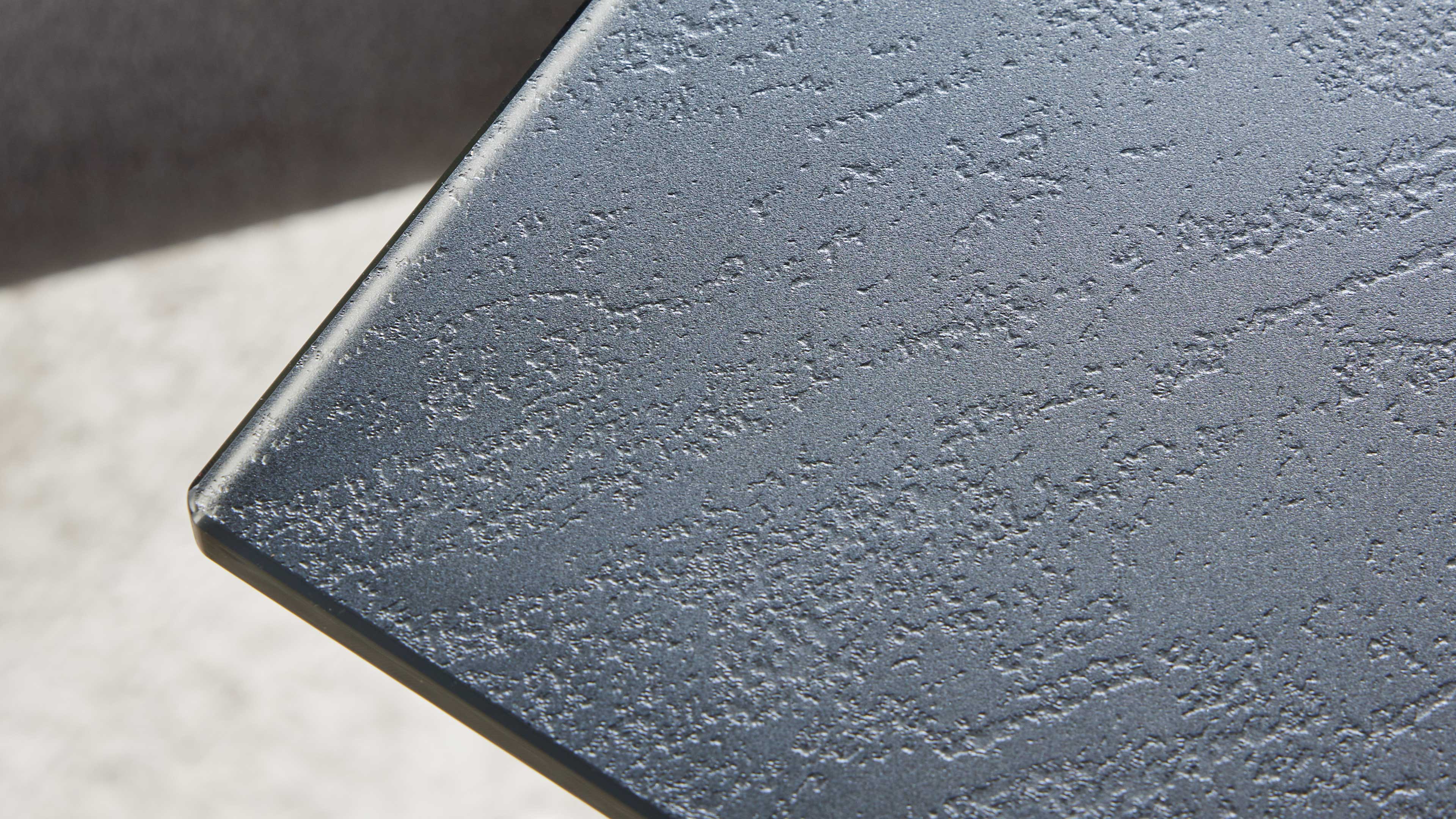 Ecke einer dunklen Tischplatte mit strukturierter, rauer Oberfläche und abgeschrägter Kante vor hellem, unscharfem Hintergrund.