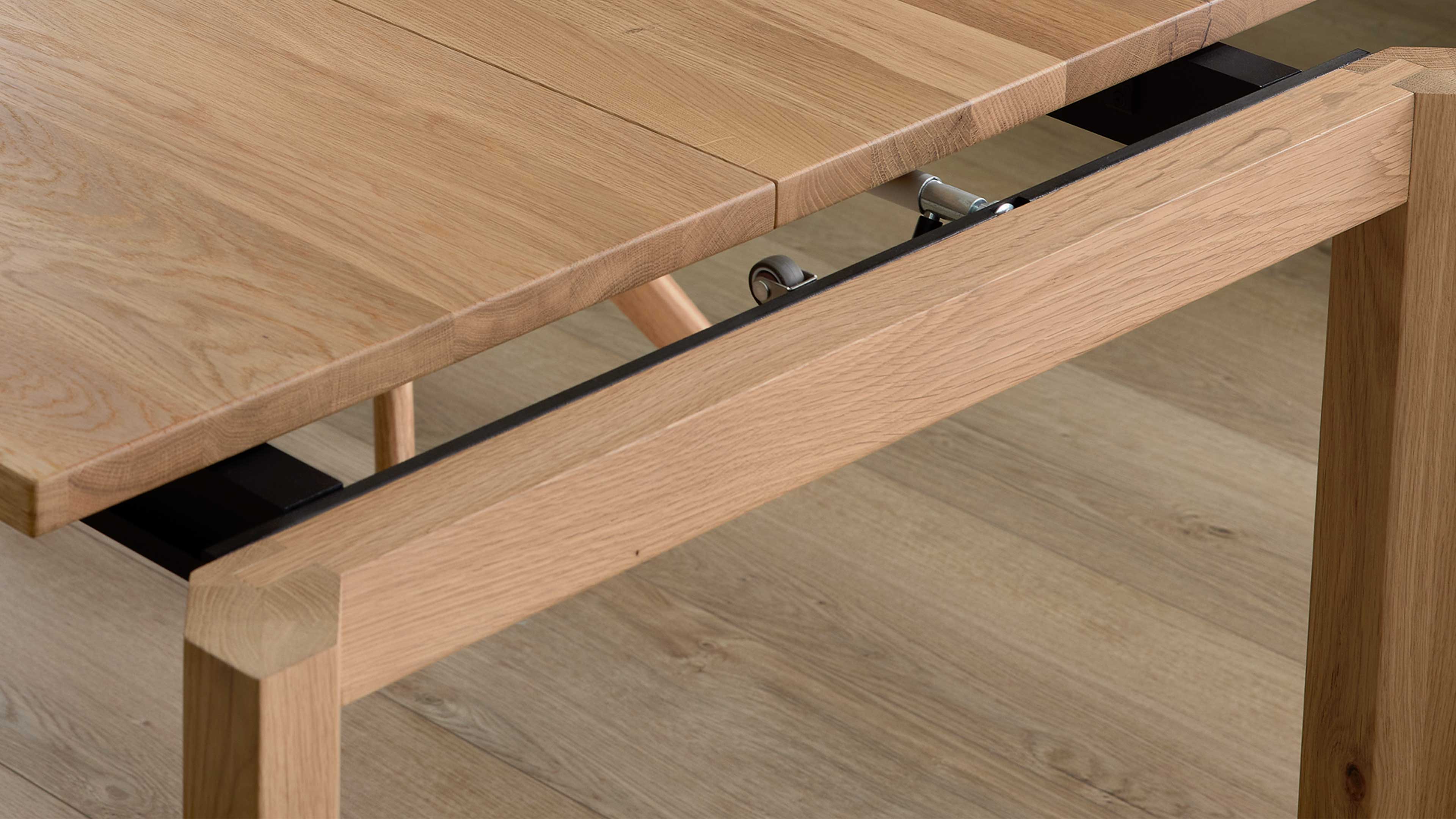 Teilweise ausgezogene Tischplatte mit sichtbarer Metallauszugsführung und Rollen, helle Eichenoberfläche und massives Tischbein.