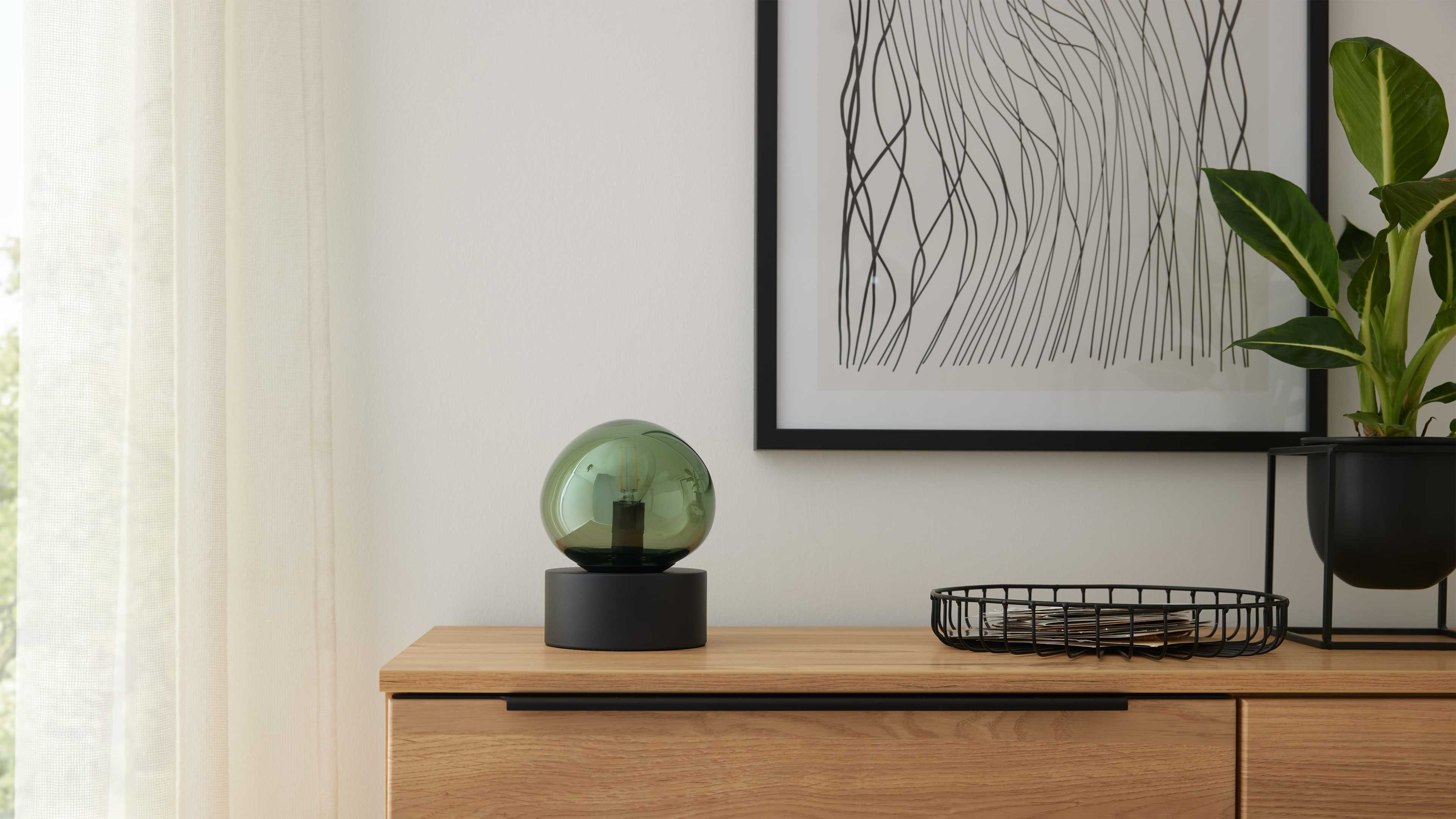Helle Holzkommode mit grüner Glaskugel-Lampe auf schwarzem Sockel, Drahtkorb, gerahmter Linienzeichnung an der Wand und Topfpflanze.