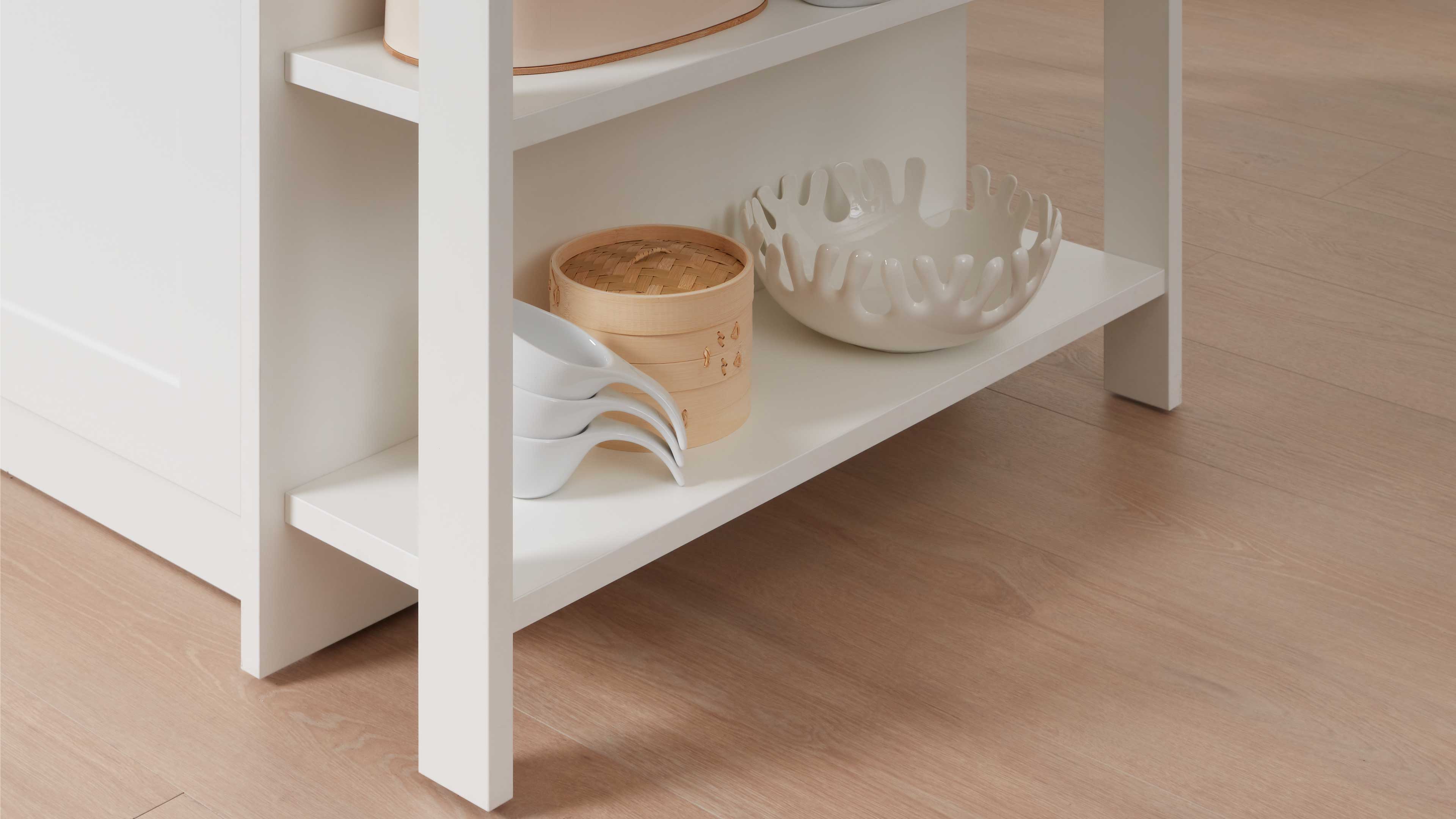 Untere Ablage eines weißen Regals mit drei weißen Löffeln, rundem Bambusdämpfer und weißer Keramikschale mit unregelmäßigem Rand.