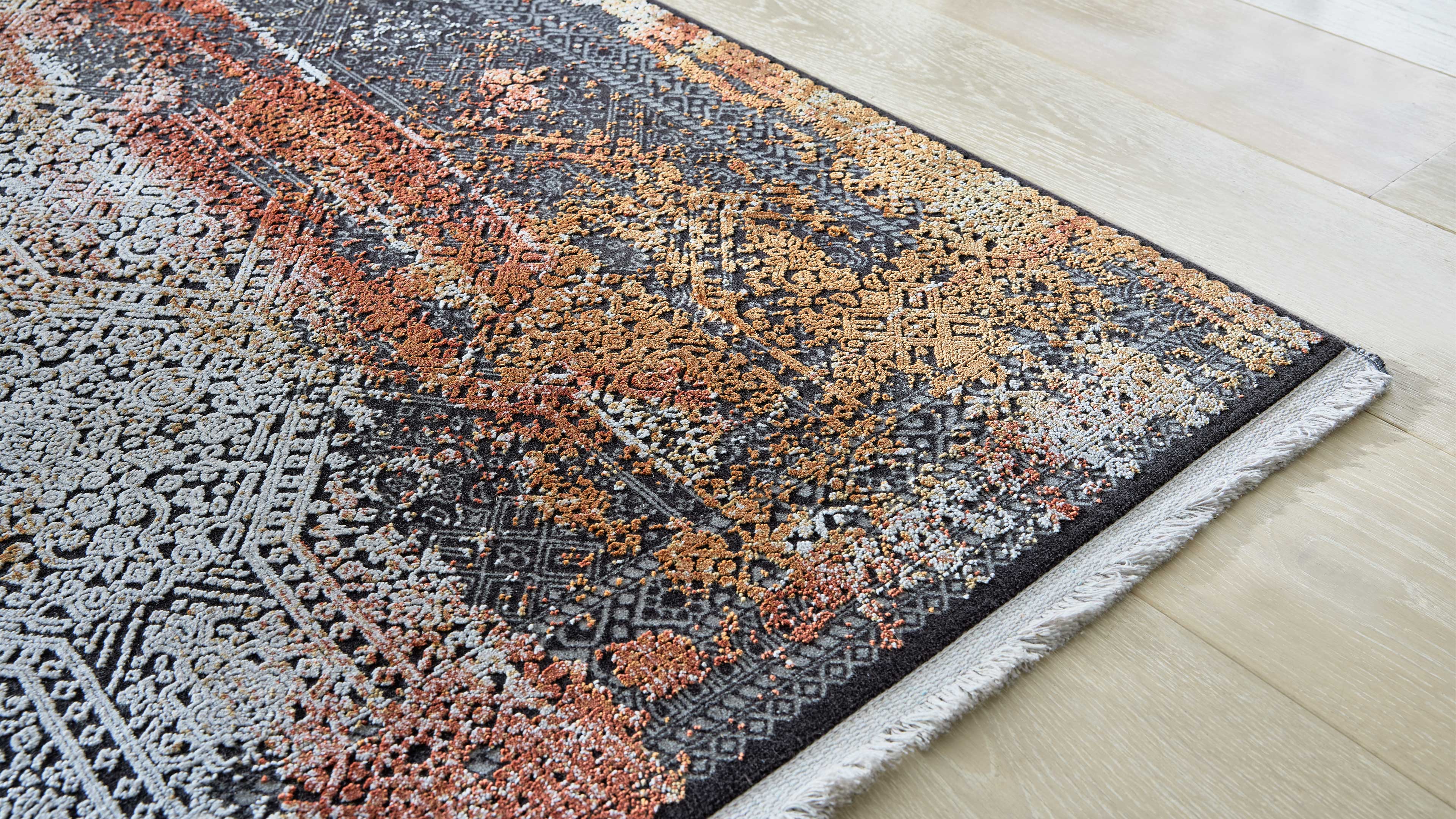 Teppichecke mit dichtem Ornamentmuster in Grau, Schwarz, Rost und Beige, gesäumt von weißer Fransenborte auf hellem Holzboden.