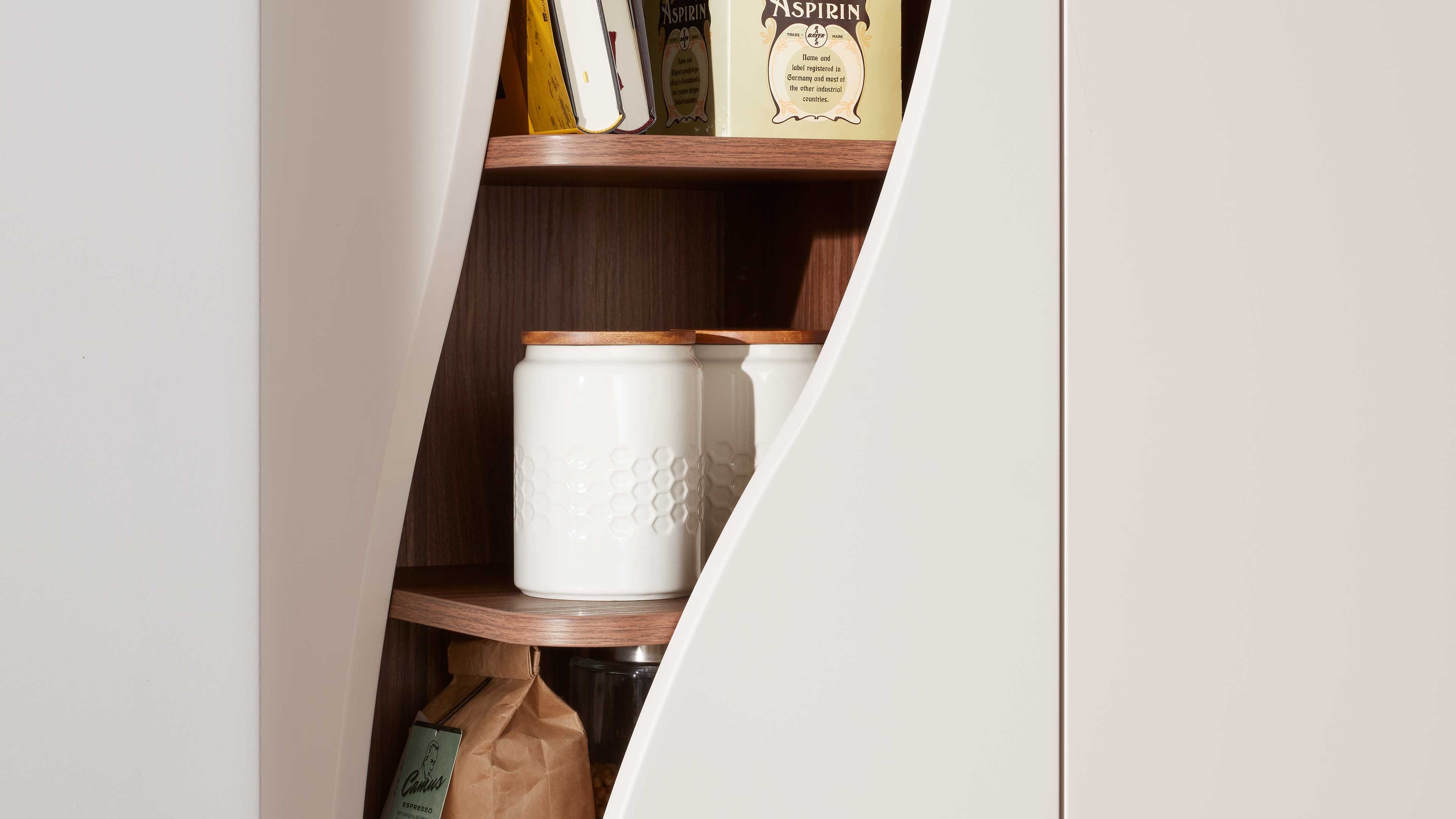 Schrank mit geschwungener, weißer Türöffnung, innen dunkle Holzregale mit weißen Vorratsdosen und Holzdeckeln.