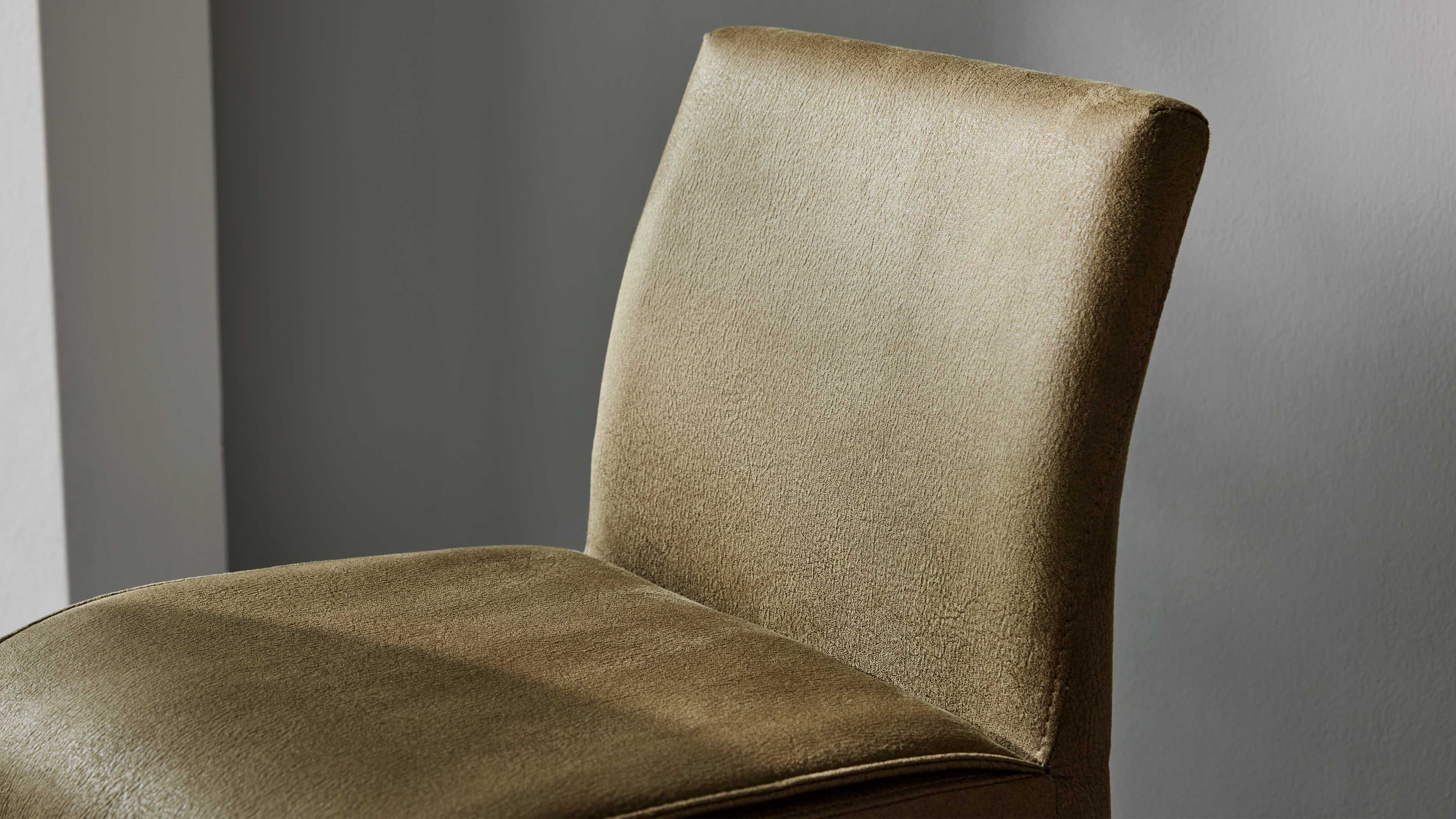 Stuhlrücken und Sitzpolster in olivgrünem, leicht schimmerndem Bezug mit sichtbarer Naht und gerundeter Rückenlehne vor grauer Wand.