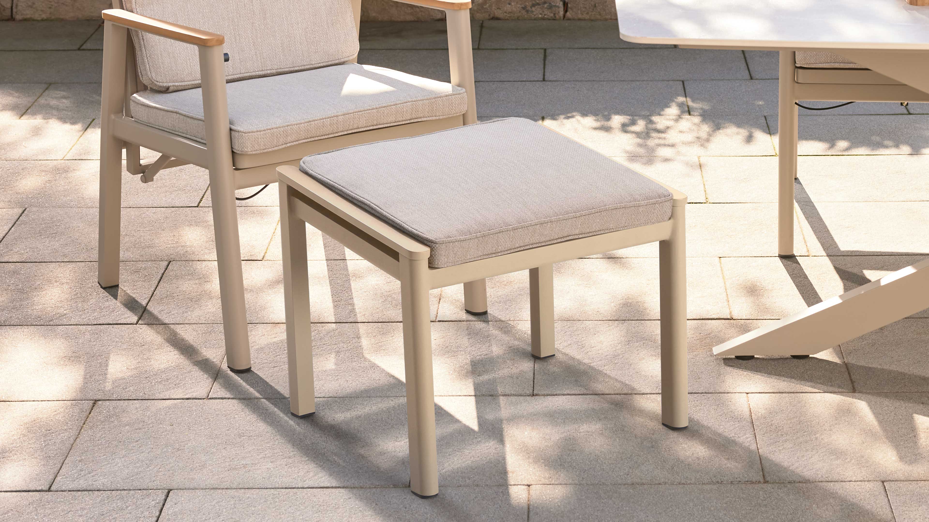 Beiger quadratischer Hocker mit hellgrauem Polsterkissen auf gepflasterter Terrasse, links Stuhl, rechts Tischbein.