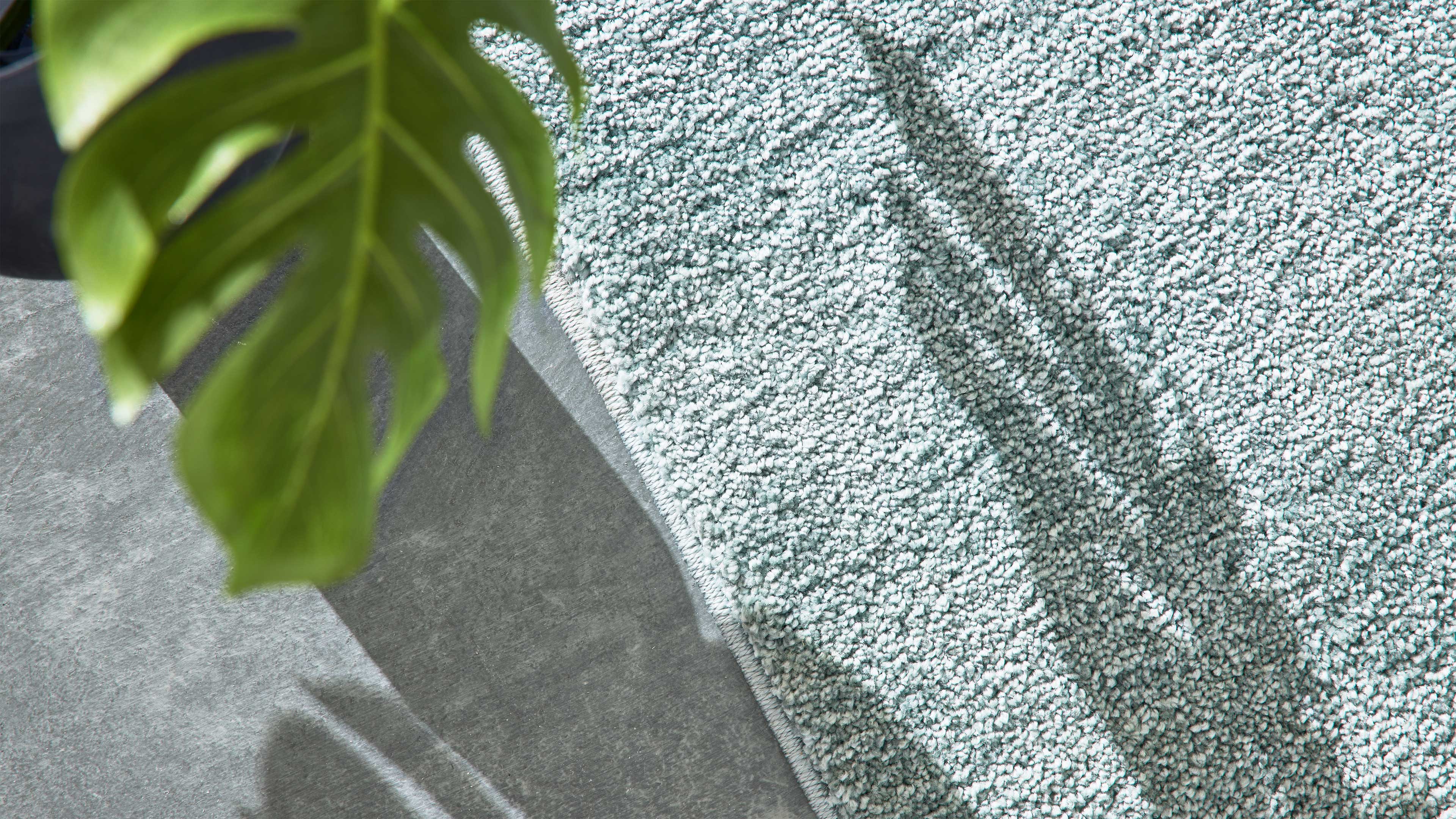 Hellmintgrüner, dicht gearbeiteter Teppich mit körniger Oberfläche liegt auf grauem Boden; links oben unscharfes grünes Pflanzenblatt.