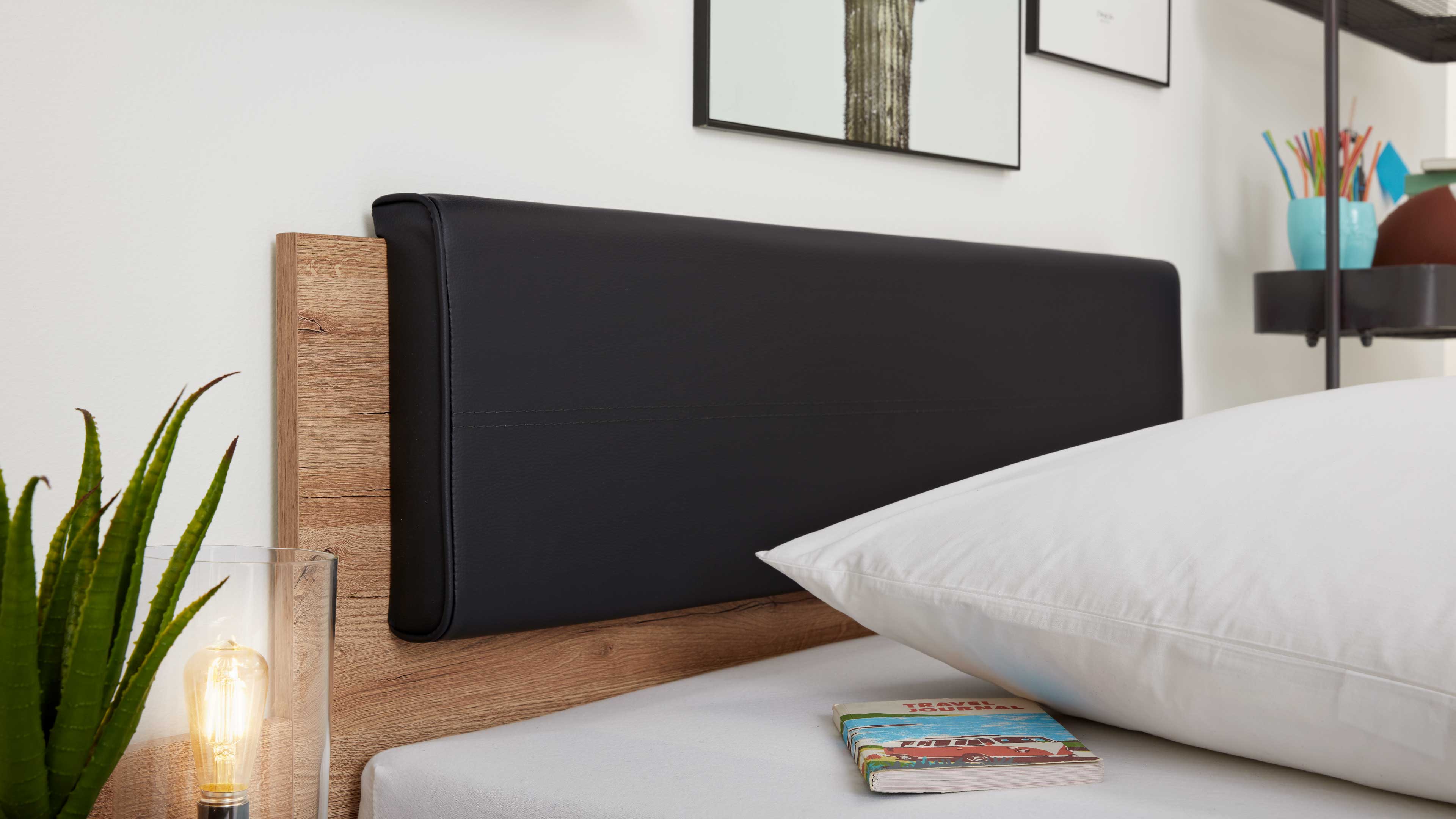 Schwarze, gepolsterte Kopfteilauflage an hellem Holzrahmen, links Pflanze und Glaslampe, rechts Kissen und Notizbuch.