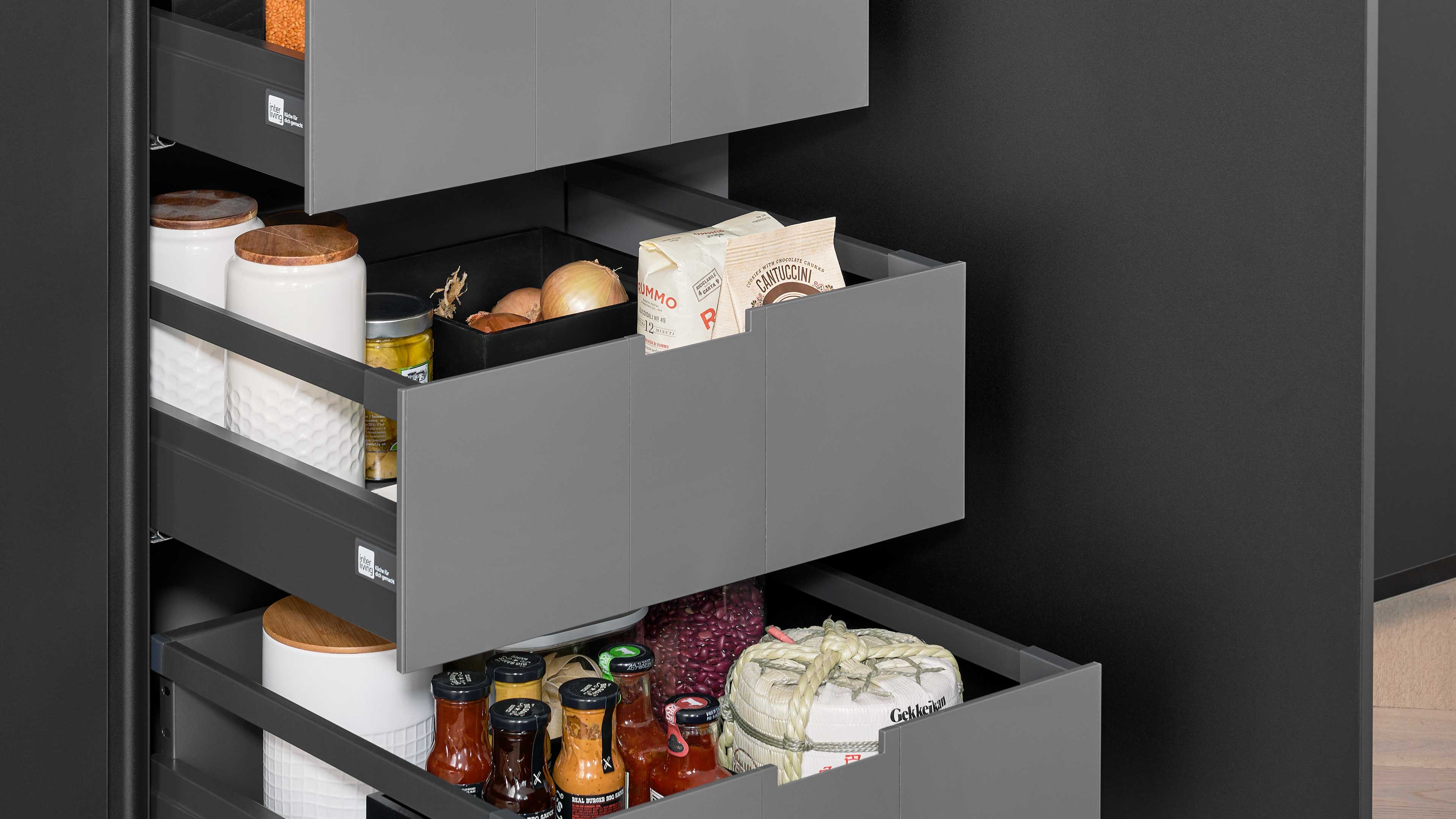 Geöffnete graue Küchenschubladen mit eingeteilten Fächern, darin Gewürzgläser, Vorratstüten, Zwiebeln und Dosen.