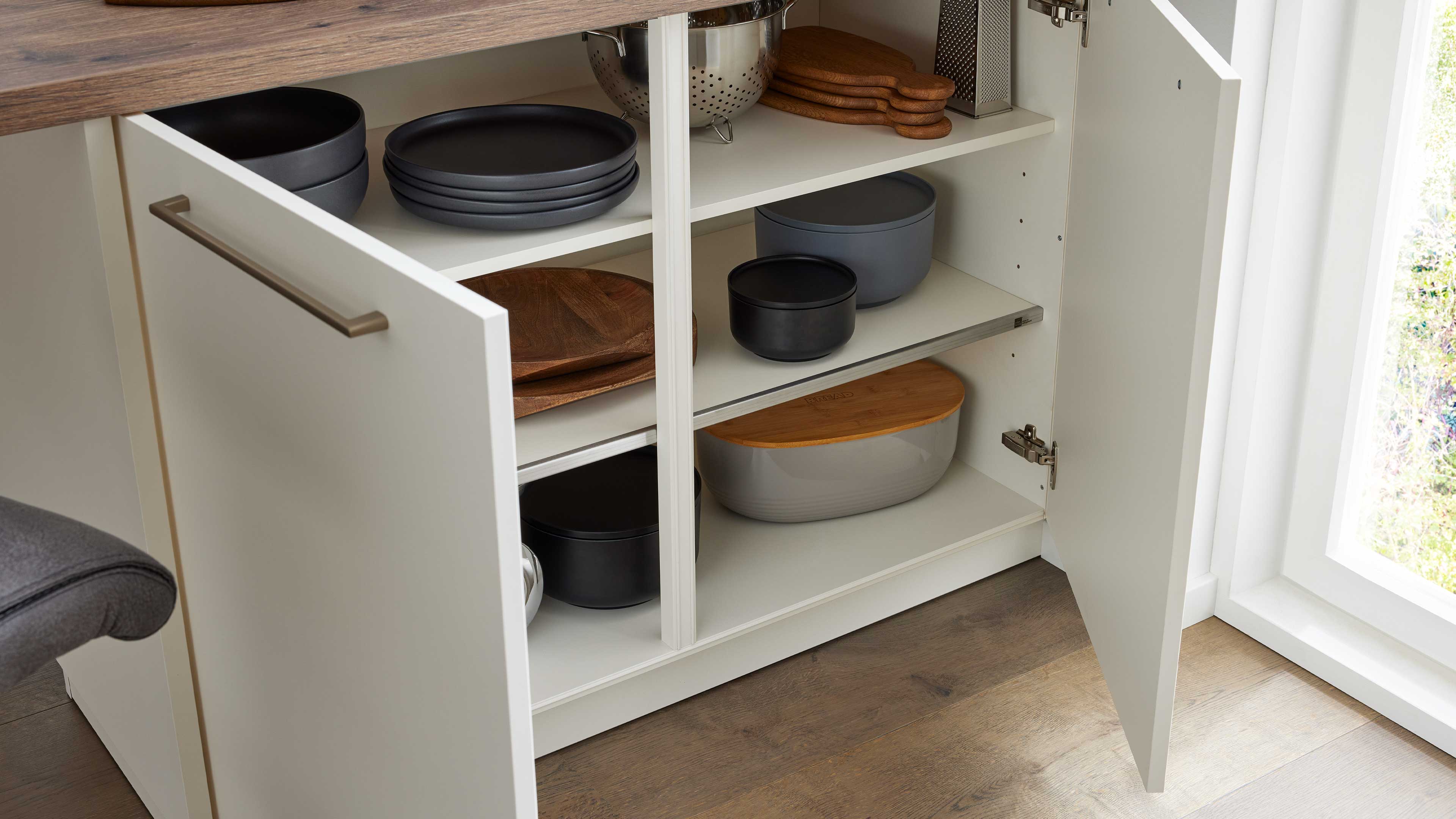 Offenes weißes Küchenschrankfach mit zwei Einlegeböden, gestapelten grauen Tellern und Schalen, Holzbrett, Edelstahlsieb und Vorratsdosen.