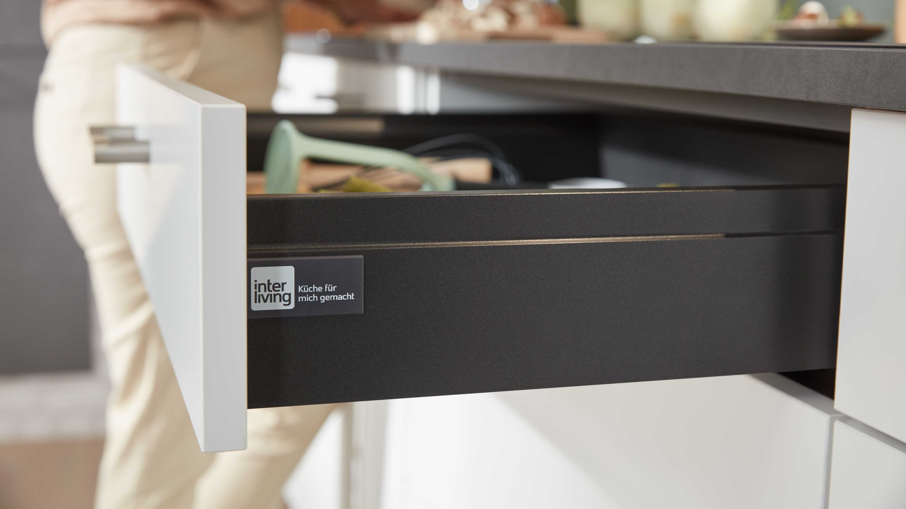 Offene Küchenschublade mit weißer Front und dunkelgrauem Innenkasten, flacher grauer Arbeitsplattenrand sichtbar.