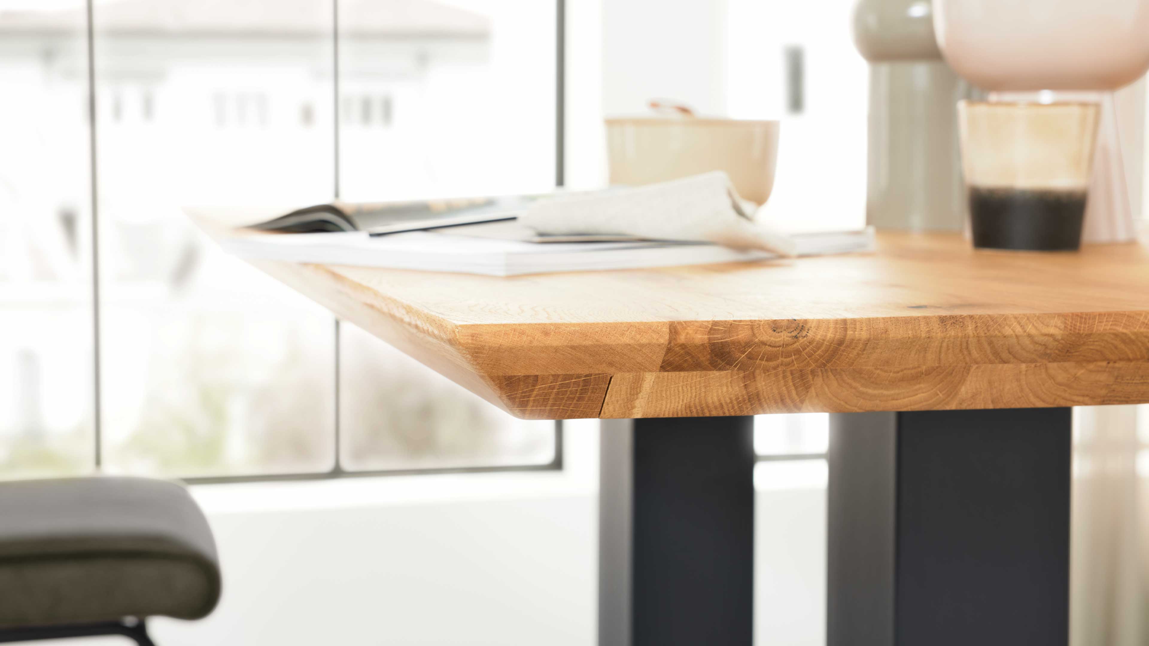 Holztischplatte mit sichtbaren Jahresringen und abgeschrägter Kante auf schwarzen Metallbeinen, darauf Glas, Magazin und Serviette.