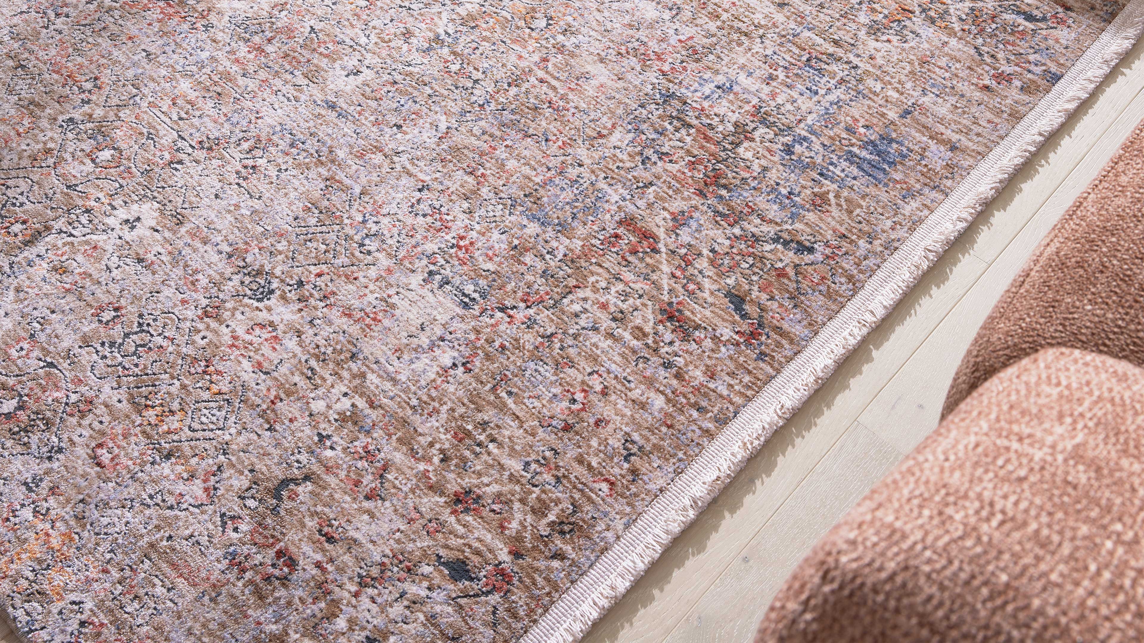 Großflächiger beige gemusterter Teppich mit Fransenrand auf hellem Holzboden, rechts Teil eines roséfarbenen Sofas sichtbar.