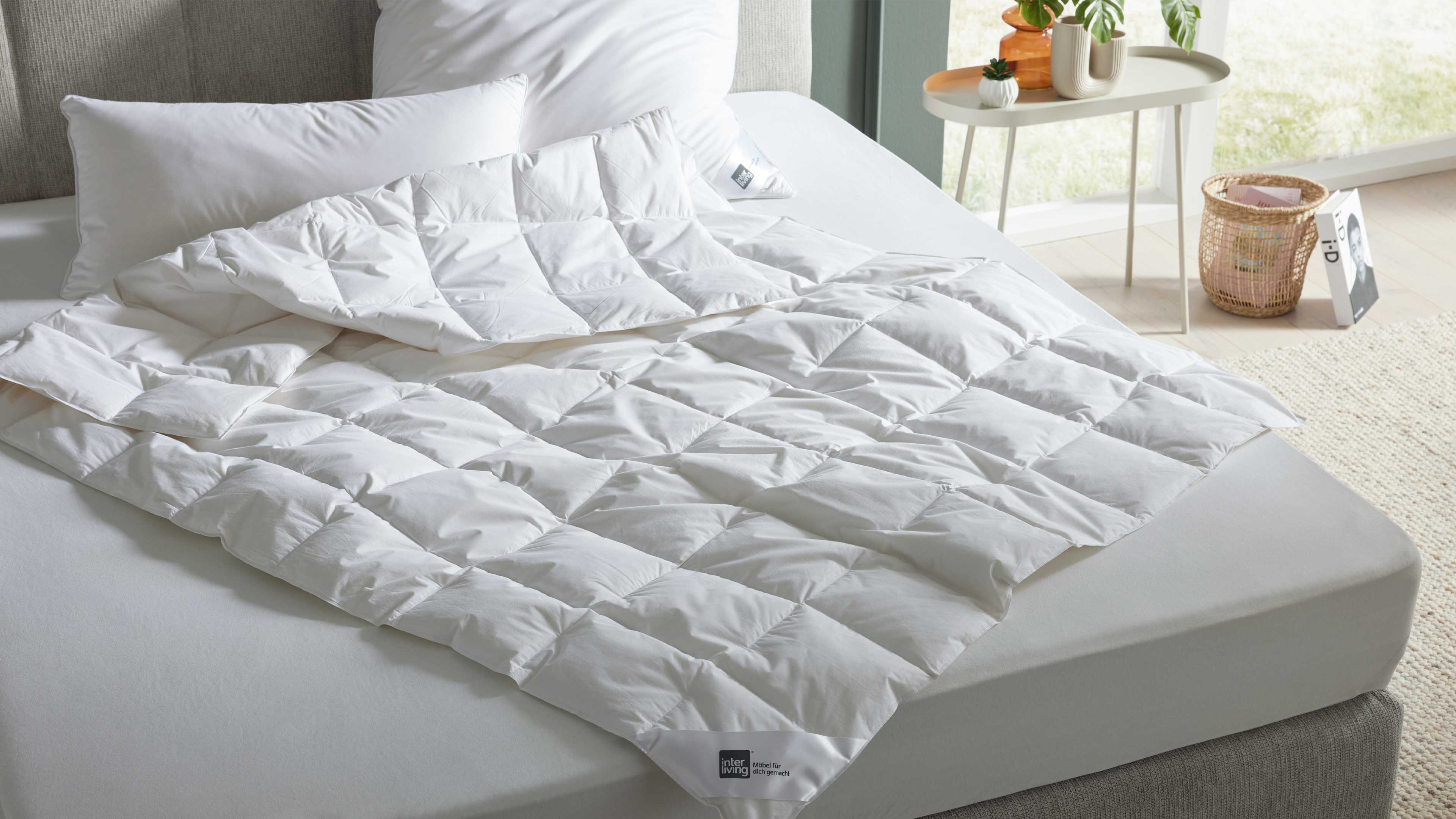Weiße gesteppte Bettdecke halb aufgefaltet auf hellgrauer Matratze zwei weiße Kissen, graues Kopfteil, Beistelltisch mit Pflanzen und Korb.