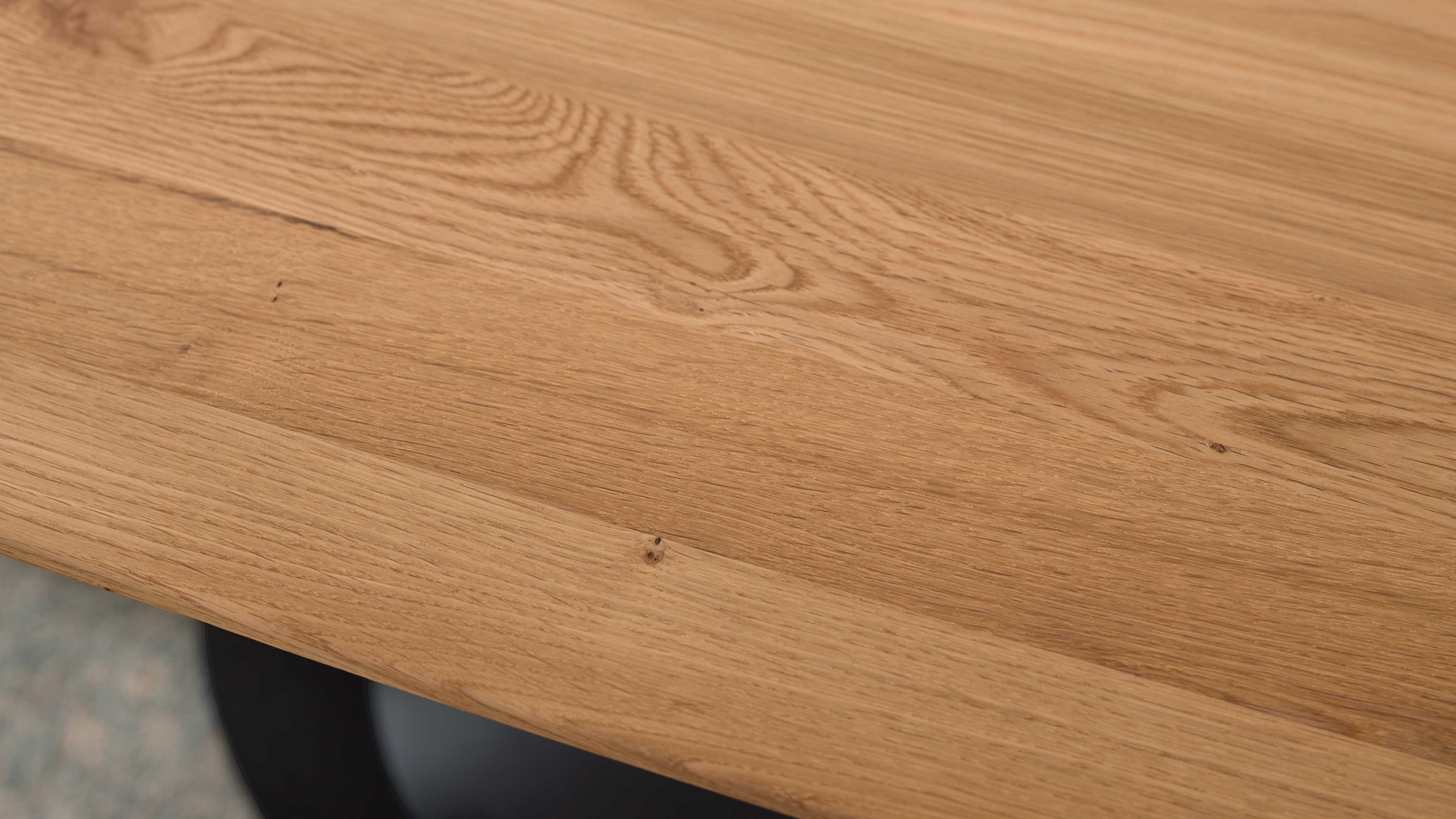 Helle Holz-Tischplatte mit sichtbarer Maserung und kleinen Ästen, am unteren Rand ein runder schwarzer Tischfußansatz.