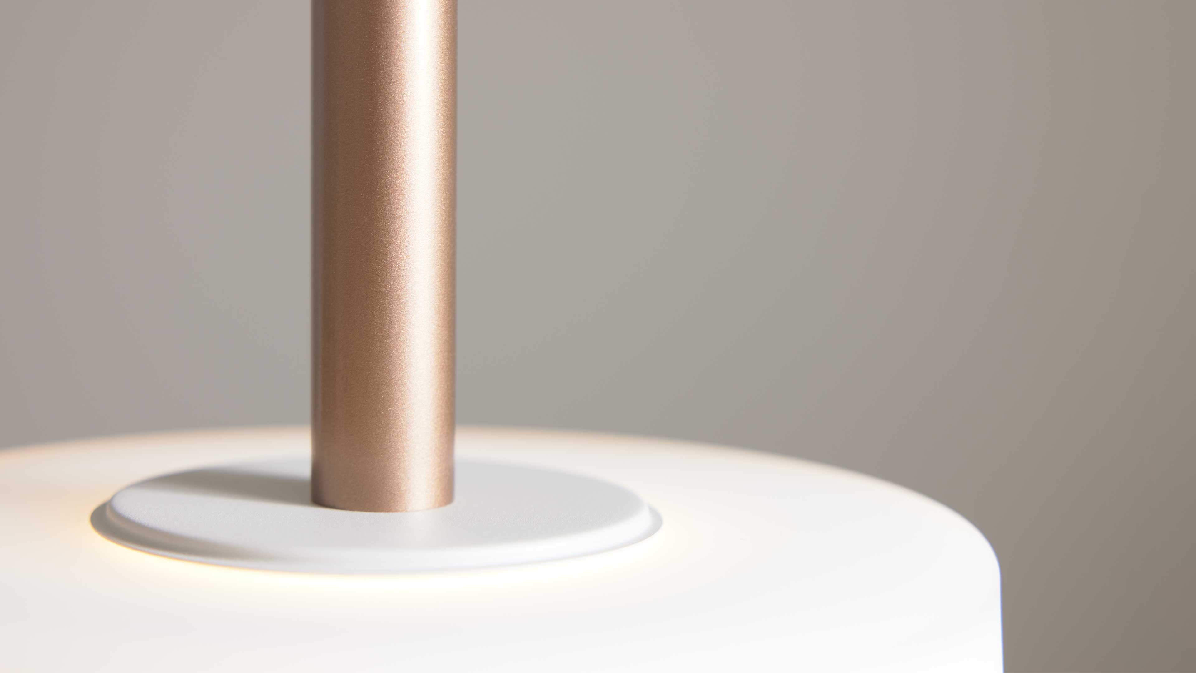 Nahaufnahme einer Lampe: goldrosafarbene Metallstange auf weißer runden Montageplatte und weißem Lampenschirm mit warmem Licht.