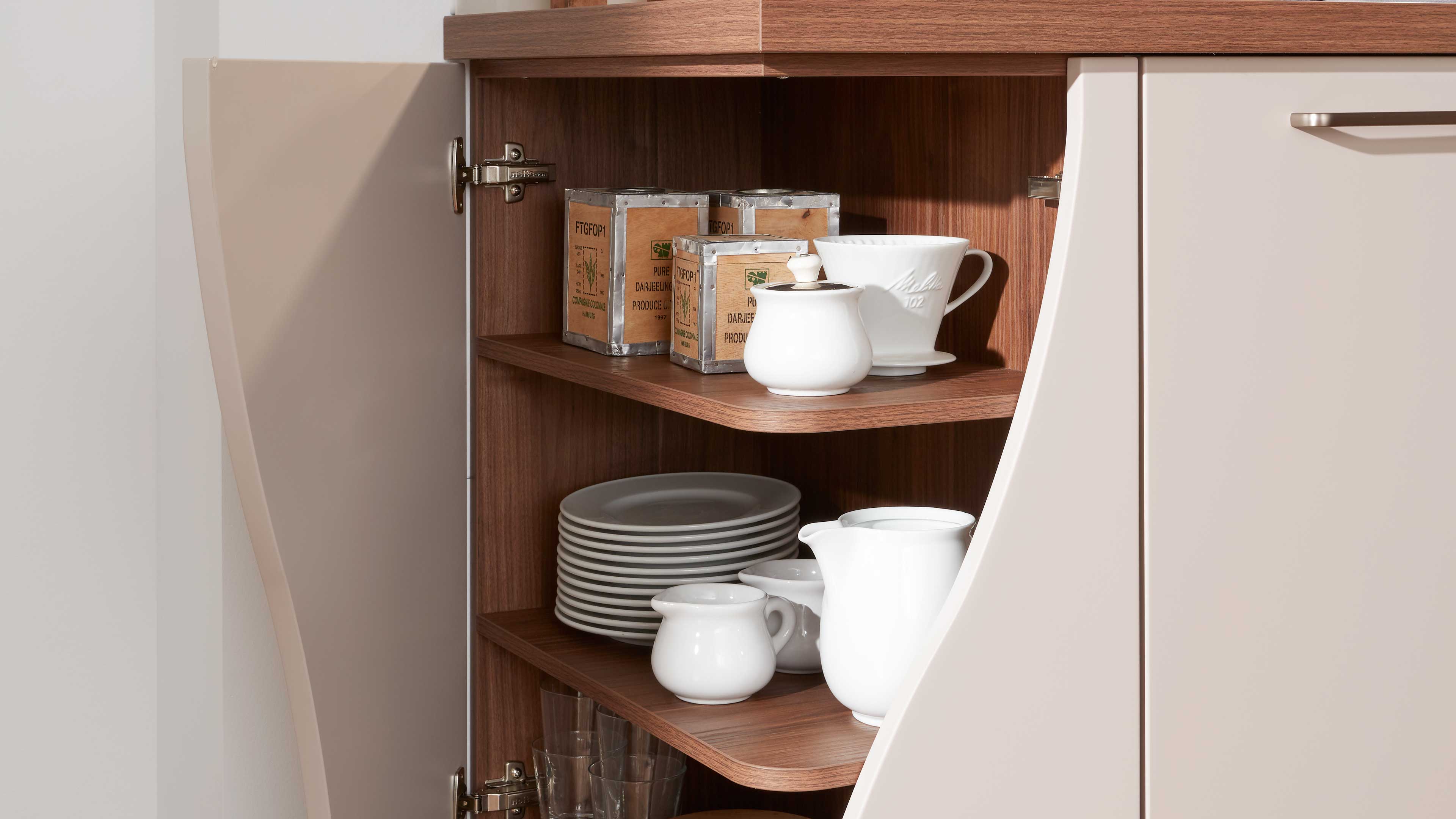 Offenes Küchen-Oberschrankfach mit warmem Holzdekor, beige Türen, weißen Keramikkannen, gestapelten Tellern und kleinen Metalldosen.