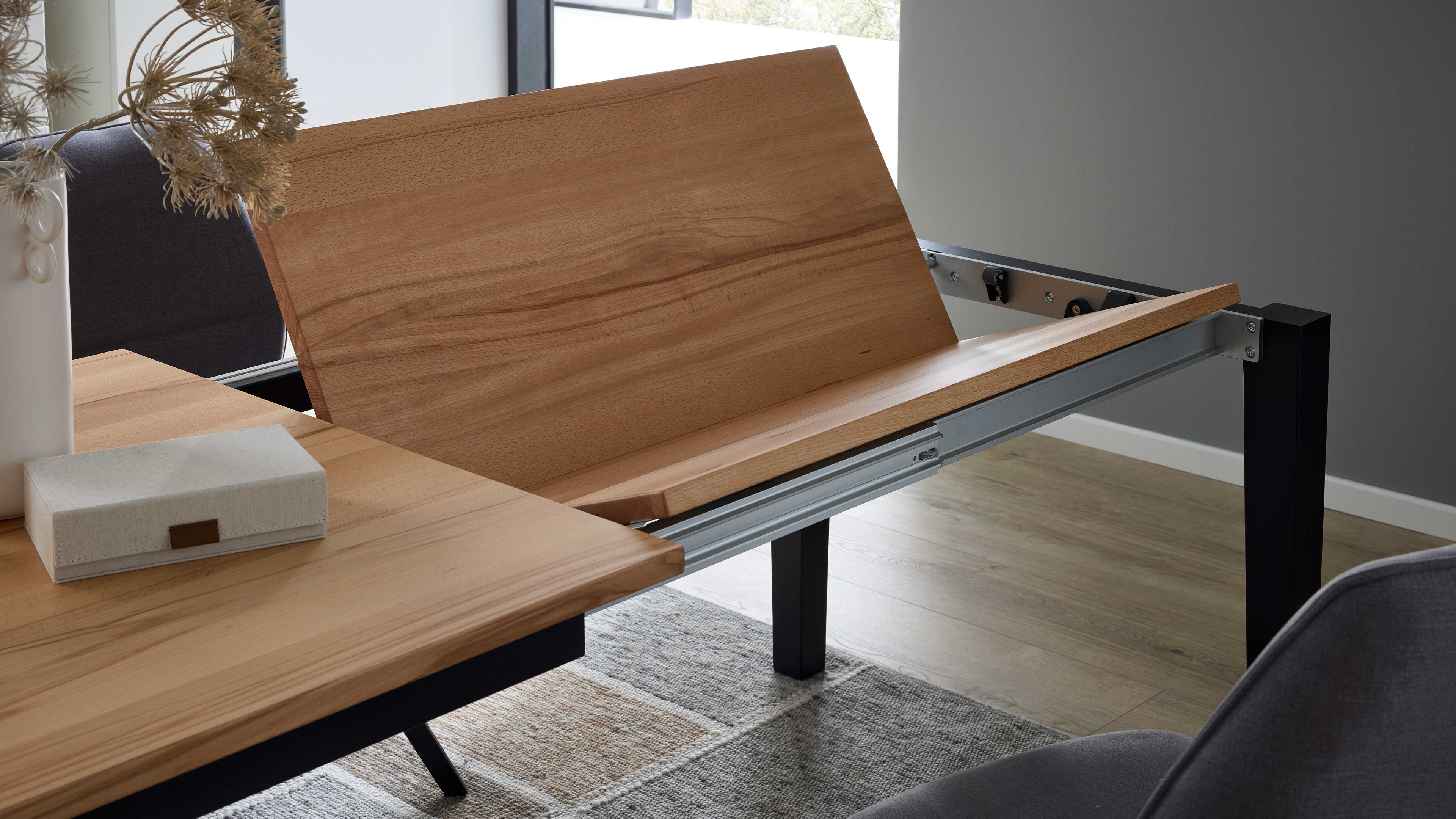 Ausgezogene Tischverlängerung aus hellbraunem Holz auf sichtbarem Metallauszug, schwarzes Tischbein, Dekobox, Vase mit Trockenblumen.