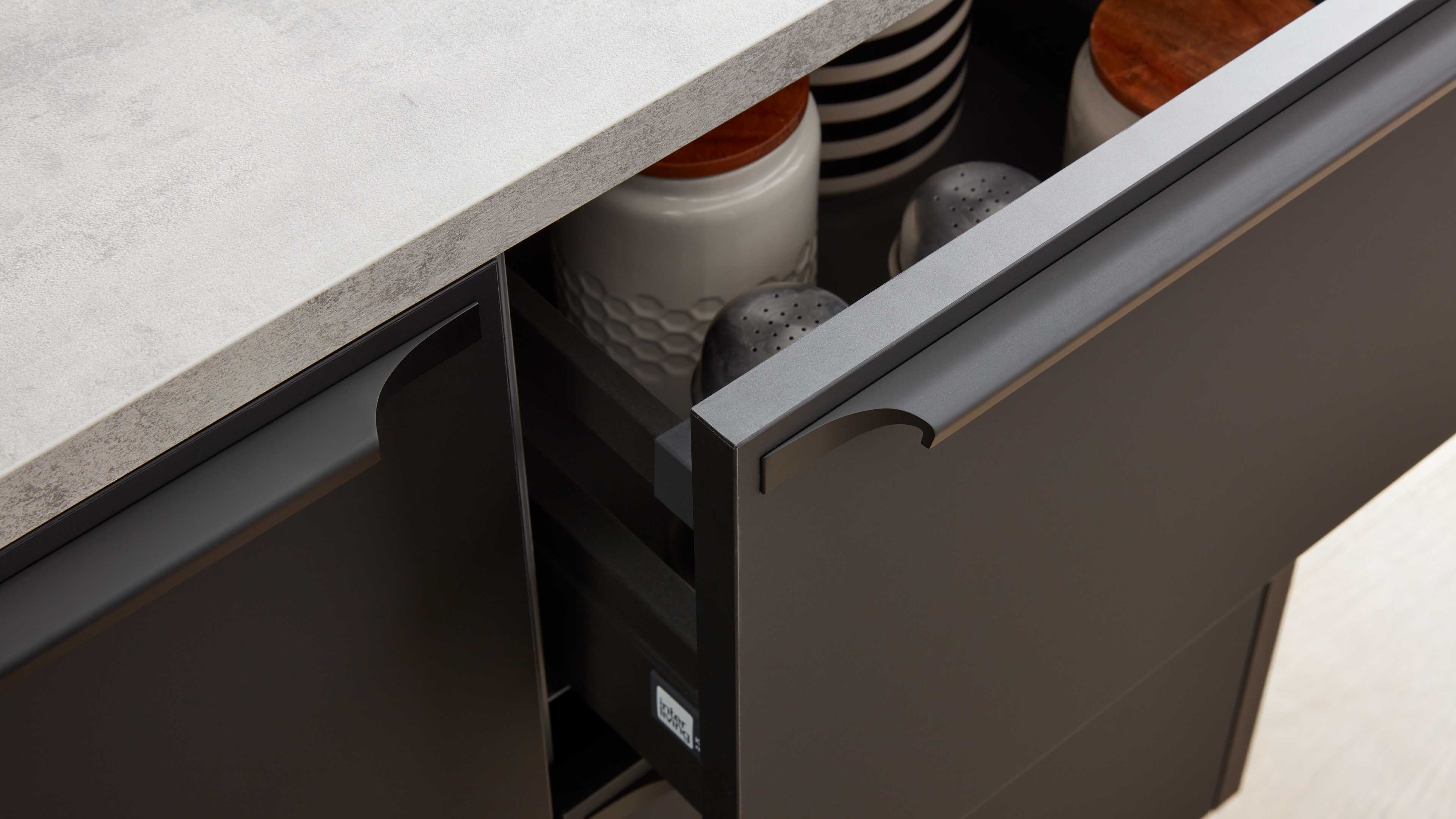 Offene dunkle Schublade unter heller Arbeitsplatte, darin weiße Dose mit Holzdeckel, zwei Metallstreuer und gestreifte Schale.