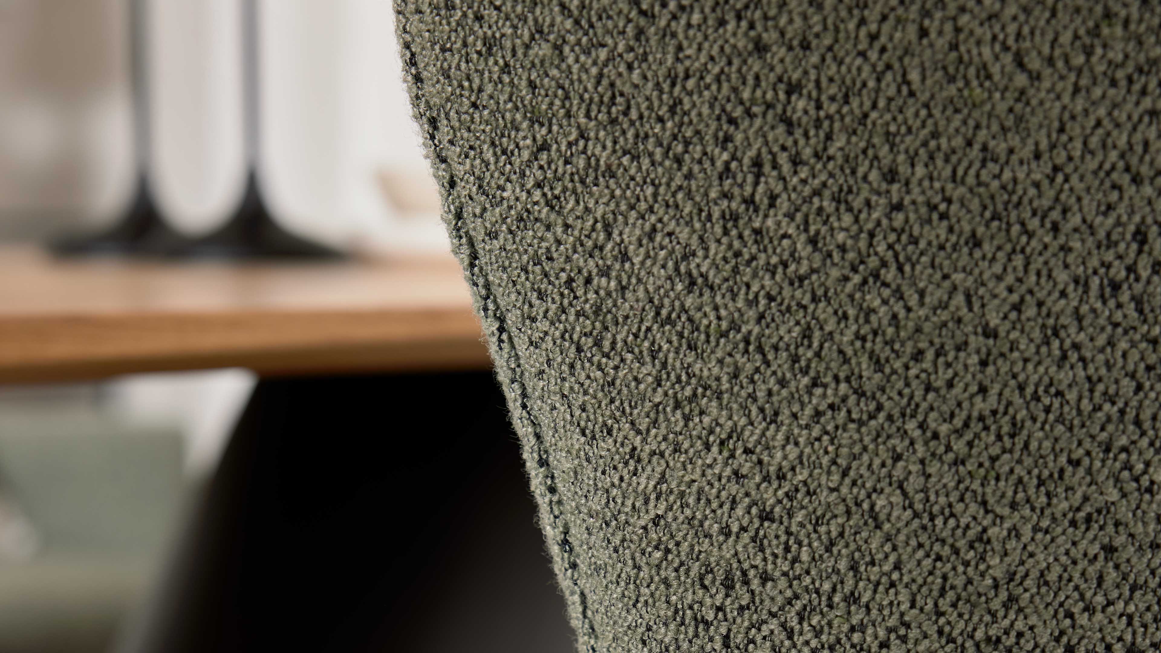 Nahaufnahme der olivegrünen, grob strukturierten Polsteroberfläche einer Stuhllehne, links unscharf Tischkante und zwei schwarze Lampenfüße.