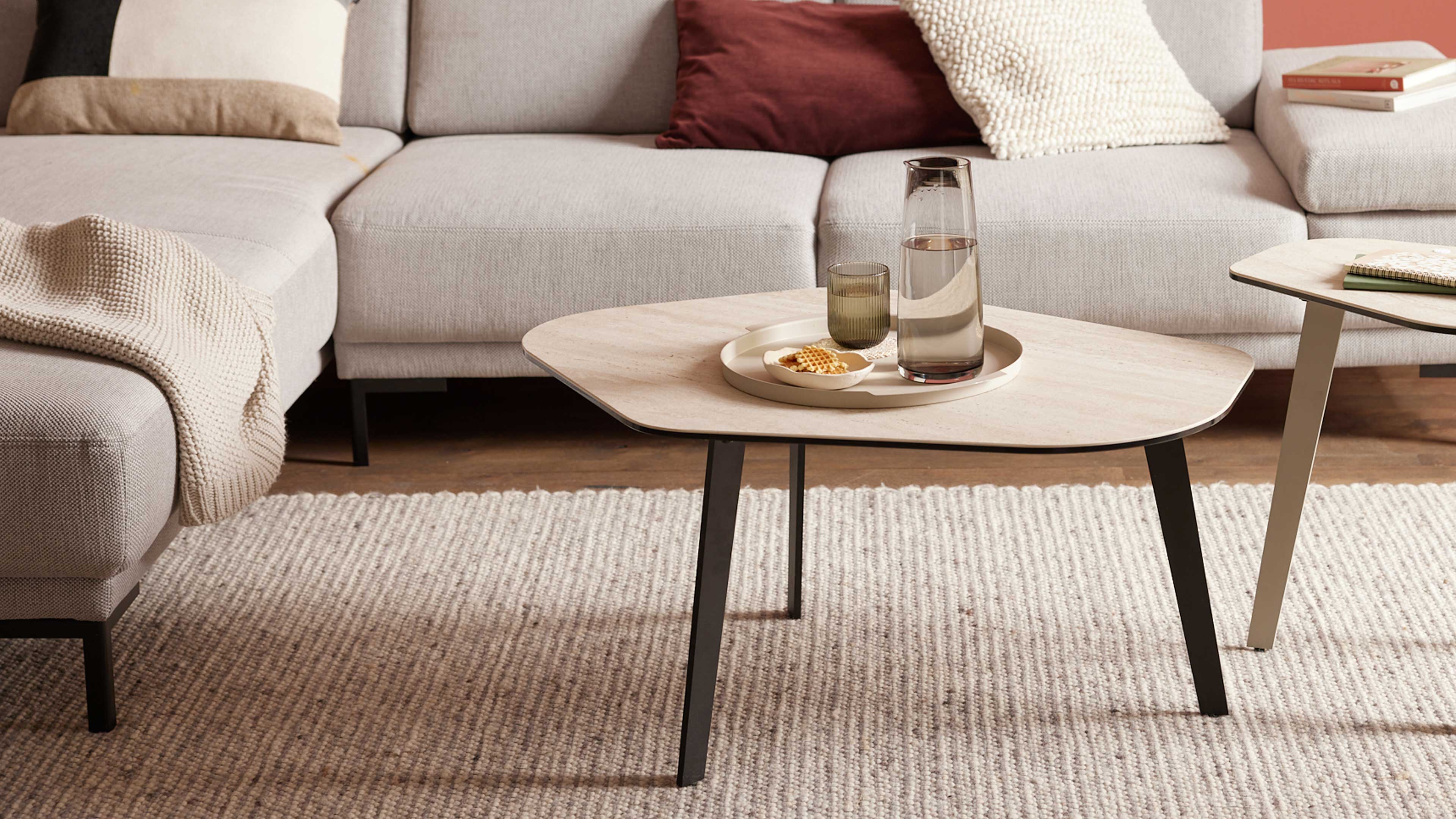 Heller Holz-Couchtisch mit schwarzem Gestell, Teller mit Karaffe, Glas und Schale vor beiger Eckcouch.